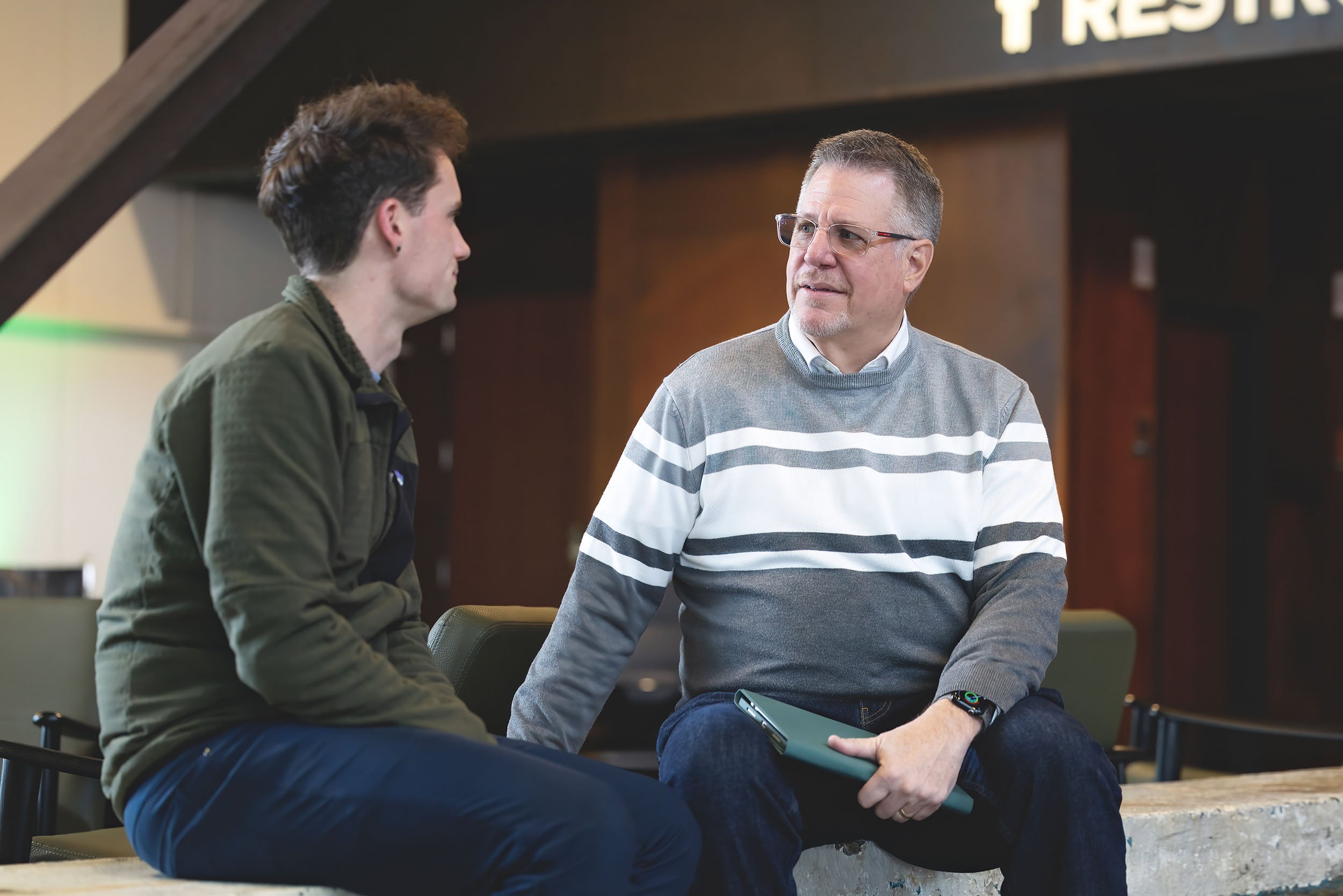 Two men sitting and talking in an indoor setting, one is older wearing glasses and a striped sweater holding a tablet, the other is younger with dark hair wearing a green jacket, behind them a sign partially visible.