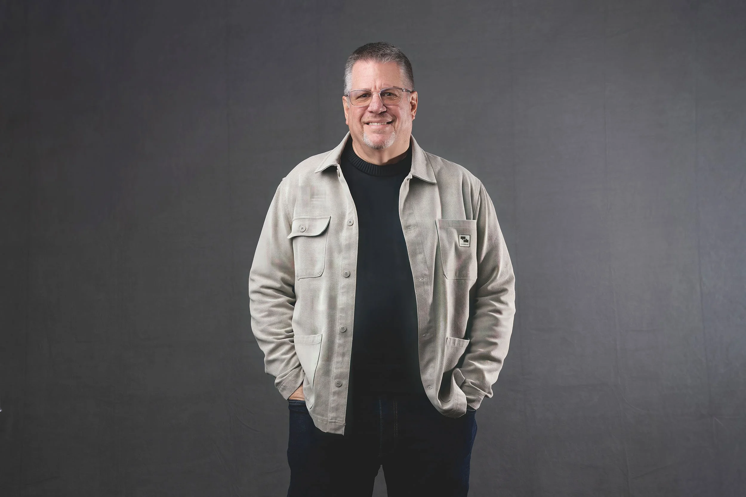 A smiling middle-aged man with short gray hair and glasses, wearing a light-colored jacket over a black shirt, standing against a gray background.