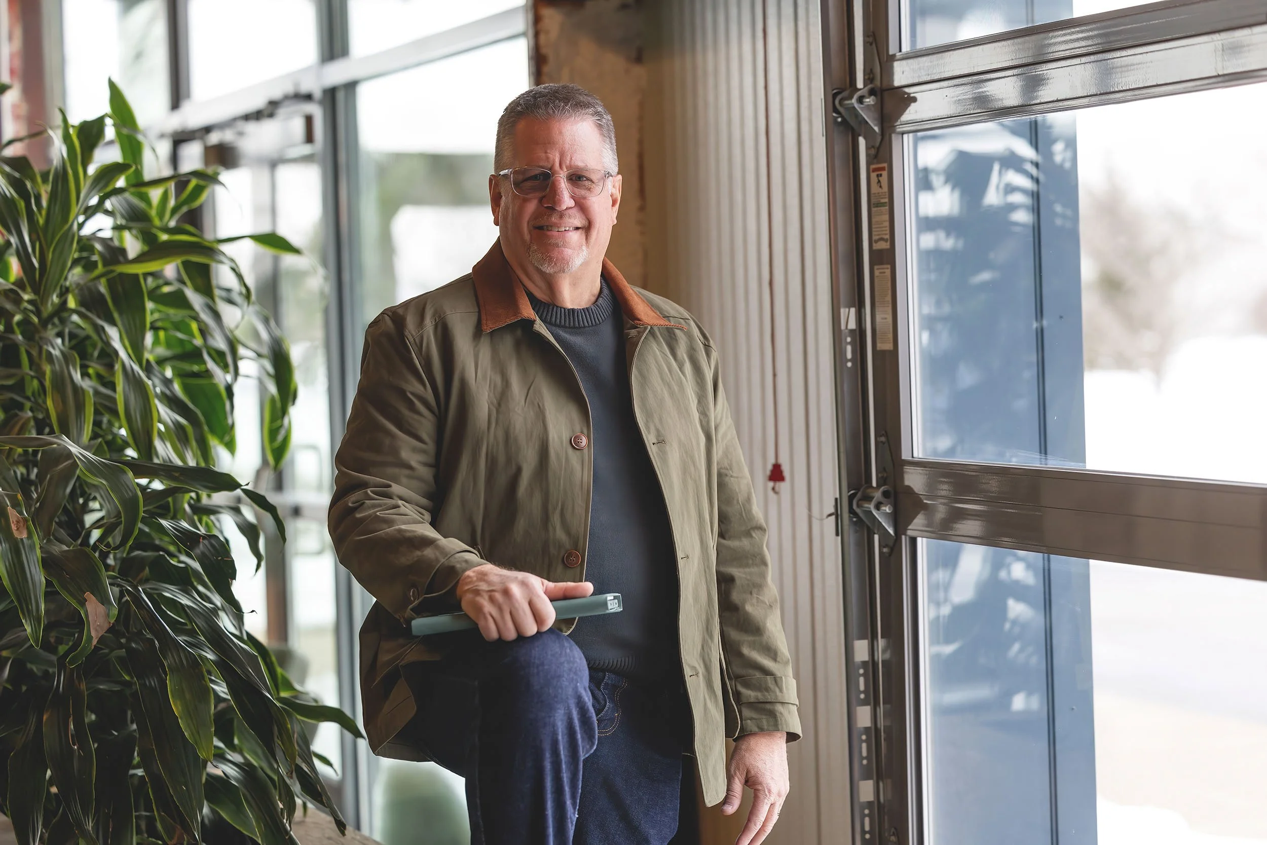 An older man with glasses, wearing a beige jacket and dark shirt, smiles while standing near a large window inside a building that appears to be a greenhouse or plant shop, holding a remote or small device in his right hand.