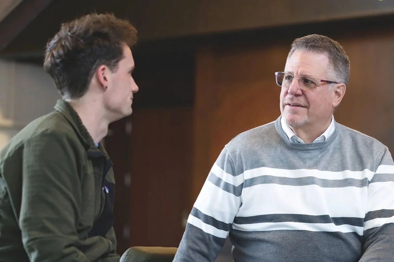 Two men talking indoors, one younger with dark hair and a green jacket, the older with glasses and a gray striped sweater.