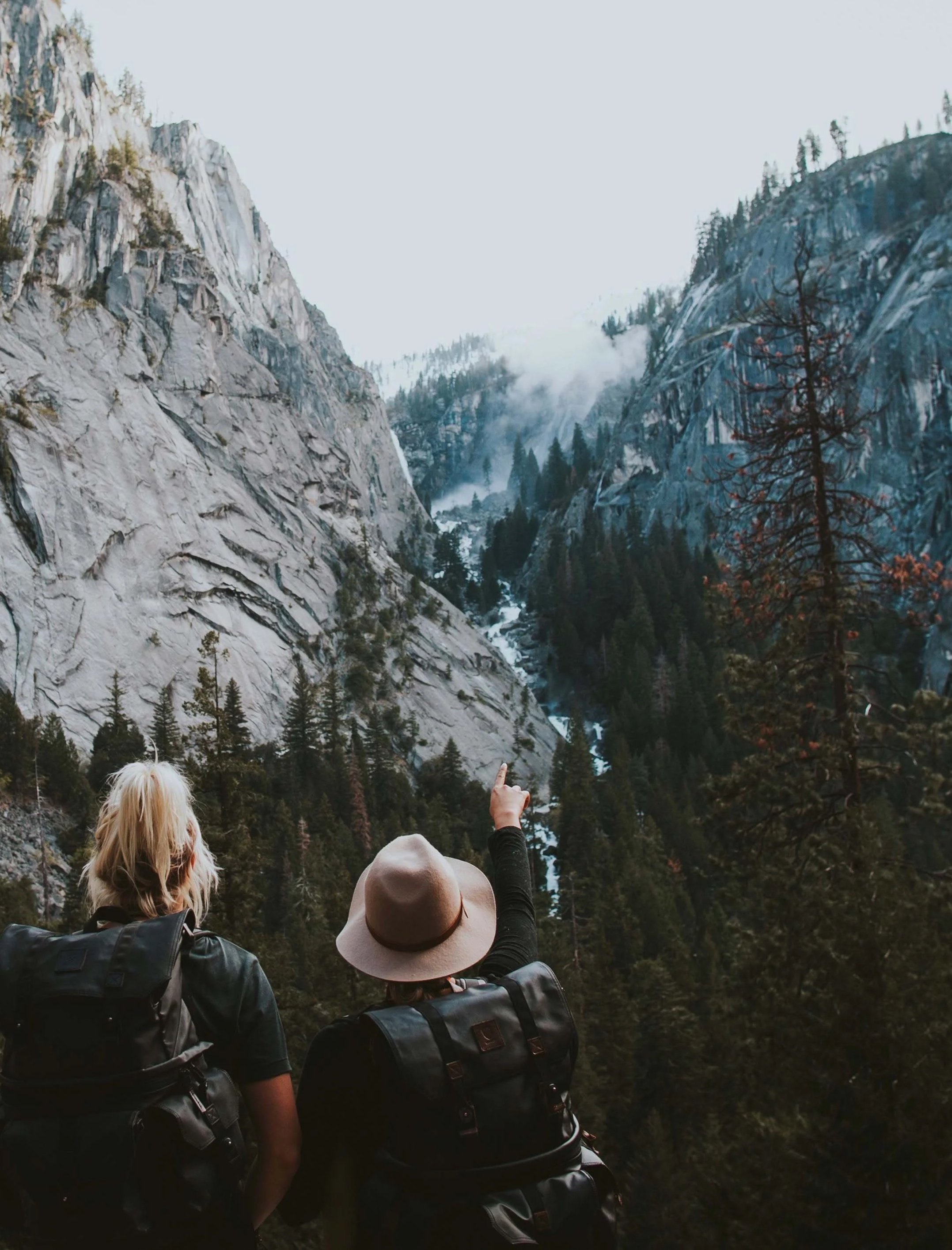 Three people with backpacks and hats looking at a mountain valley with steep cliffs and trees, some cloud and mist in the background.