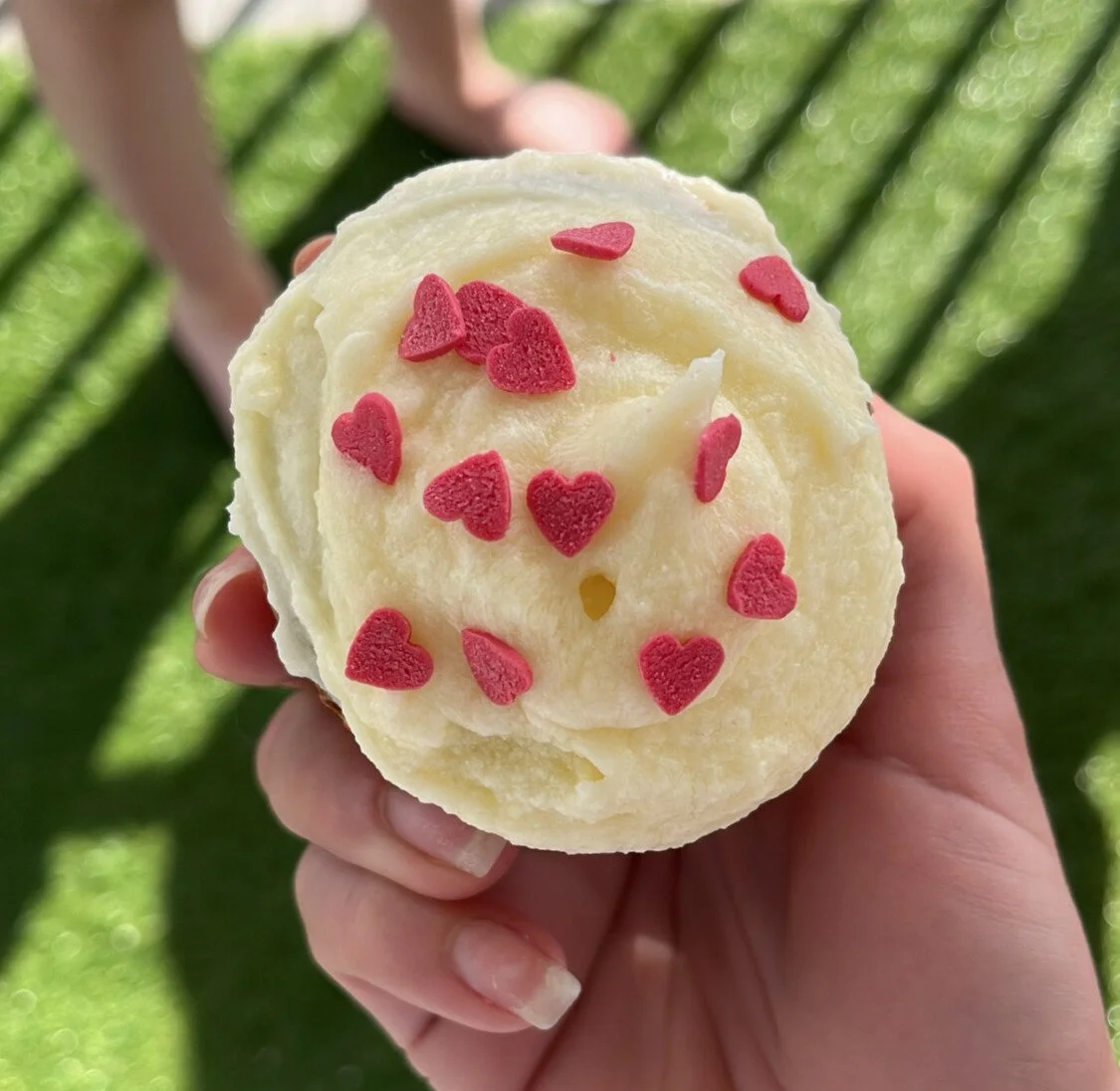 Une main tient une boule de glace à la vanille décorée de petits cœurs en bonbons rouges, avec un fond flou de jardin ou d'espace extérieur avec des plantes vertes.