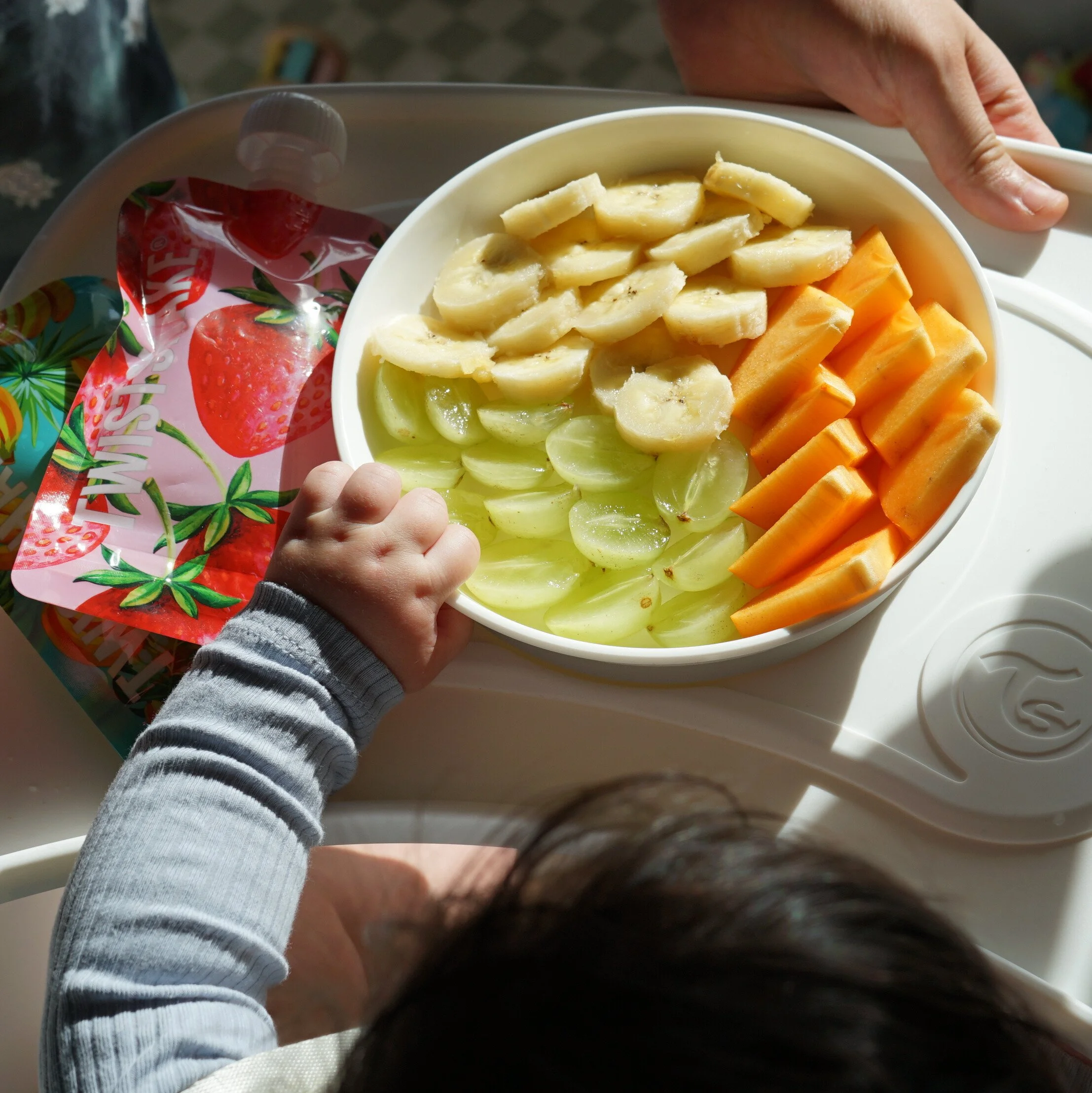 Un enfant et une adulte tenant un plateau avec un bol de fruits coupés, comprenant des bananes, des raisins verts, des morceaux de cantaloup et des morceaux de pomme.