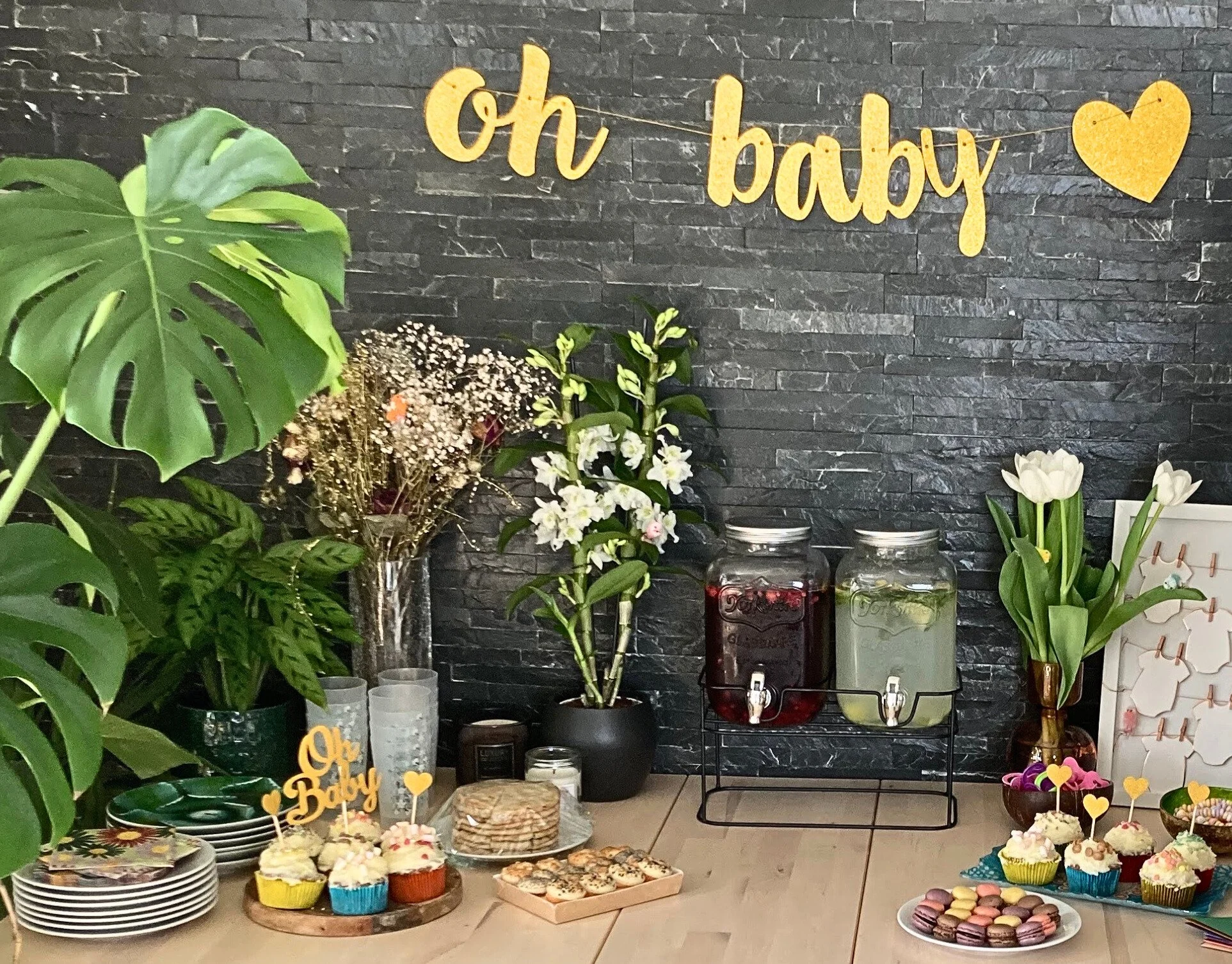 Table de fête enfant avec cupcakes, macarons, boissons, fleurs et décoration 'oh baby' sur mur noir.