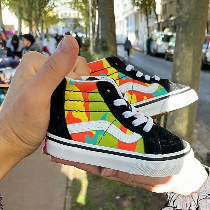 Chaussures de skateboard pour enfant avec une semelle blanche, en toile à motif coloré avec des dessins abstraits, tenues par deux mains à une rue arborée en arrière-plan.