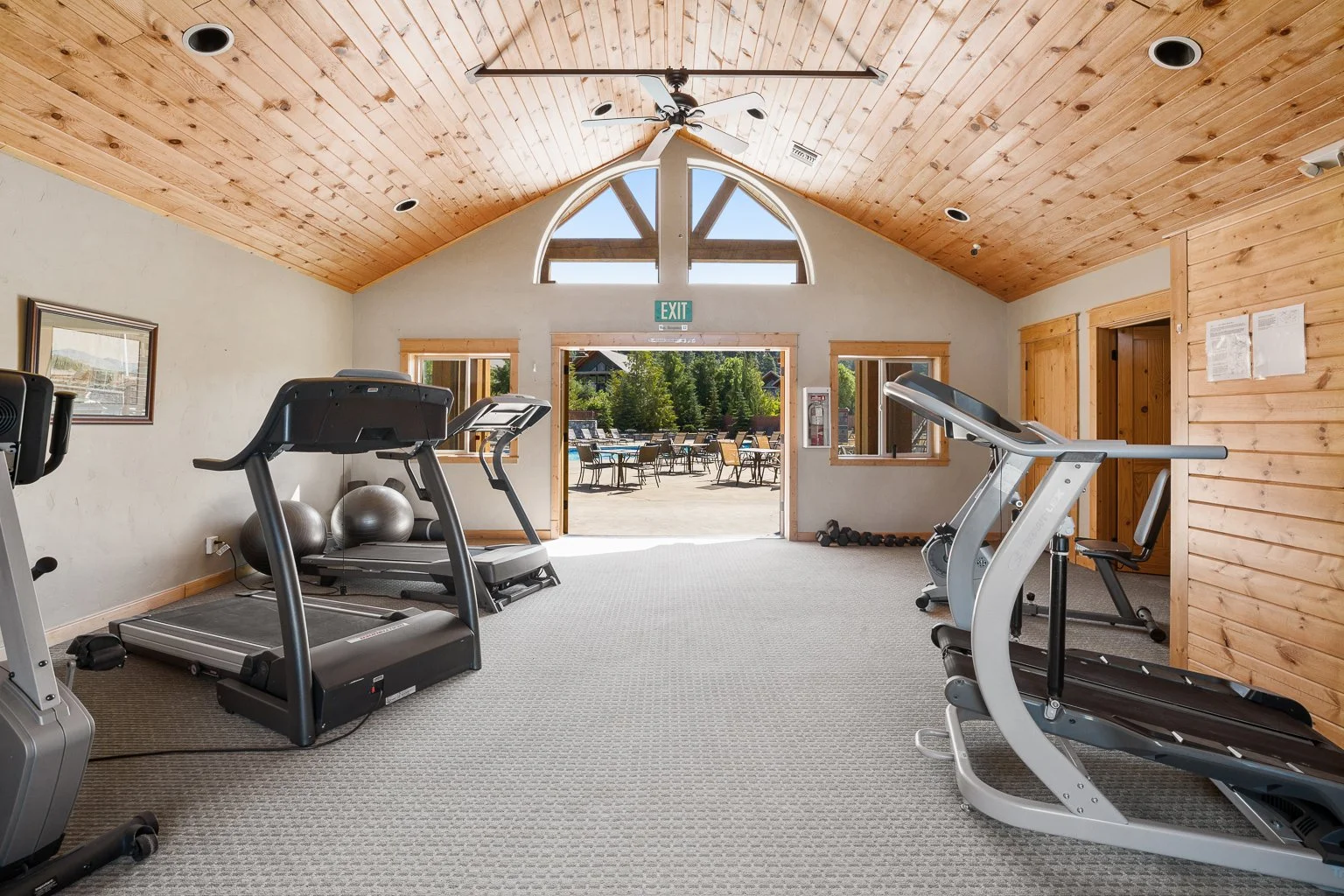 Indoor gym with treadmills and exercise equipment, wooden ceiling, open doorway leading to outdoor patio with tables and chairs, outside greenery.