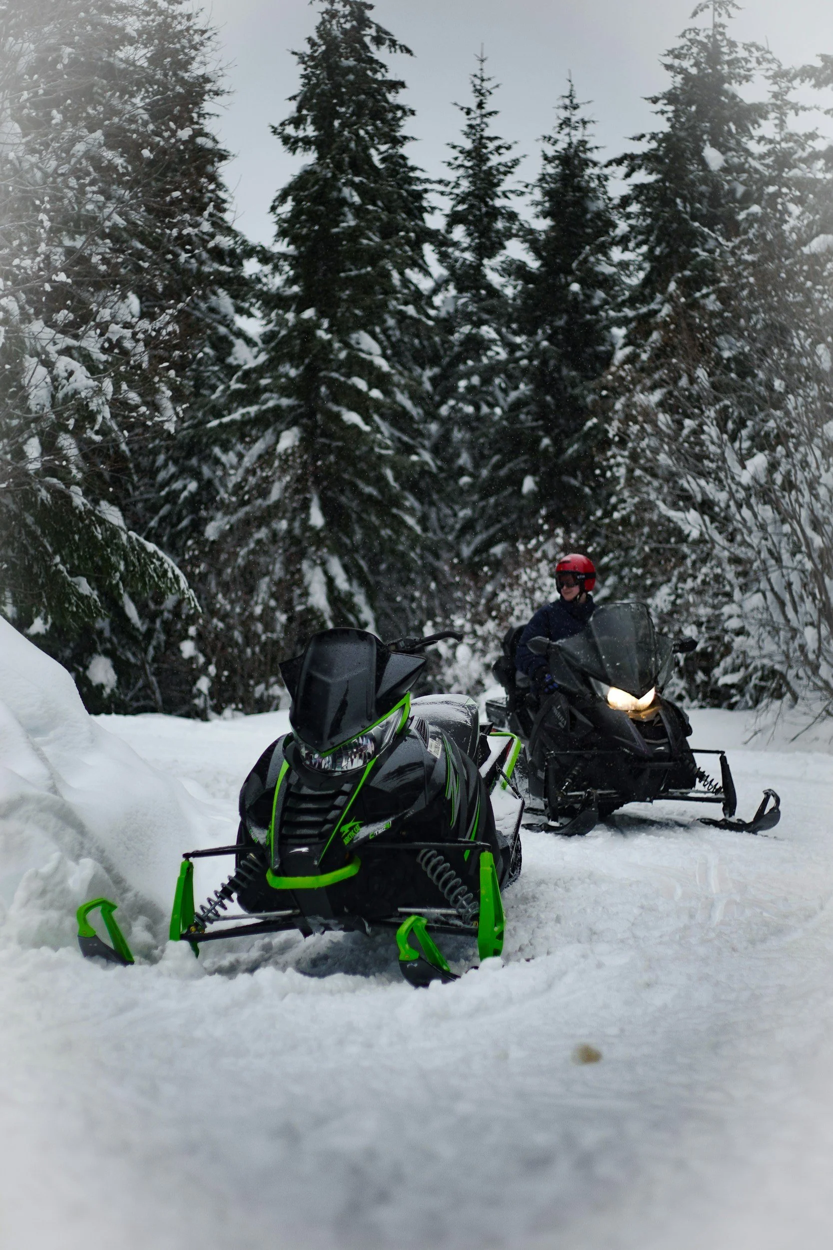 A person wearing a red helmet and dark jacket riding a snowmobile through a snowy forest. There is another snowmobile with a black and green design in the foreground. Snow-covered trees surround the scene.