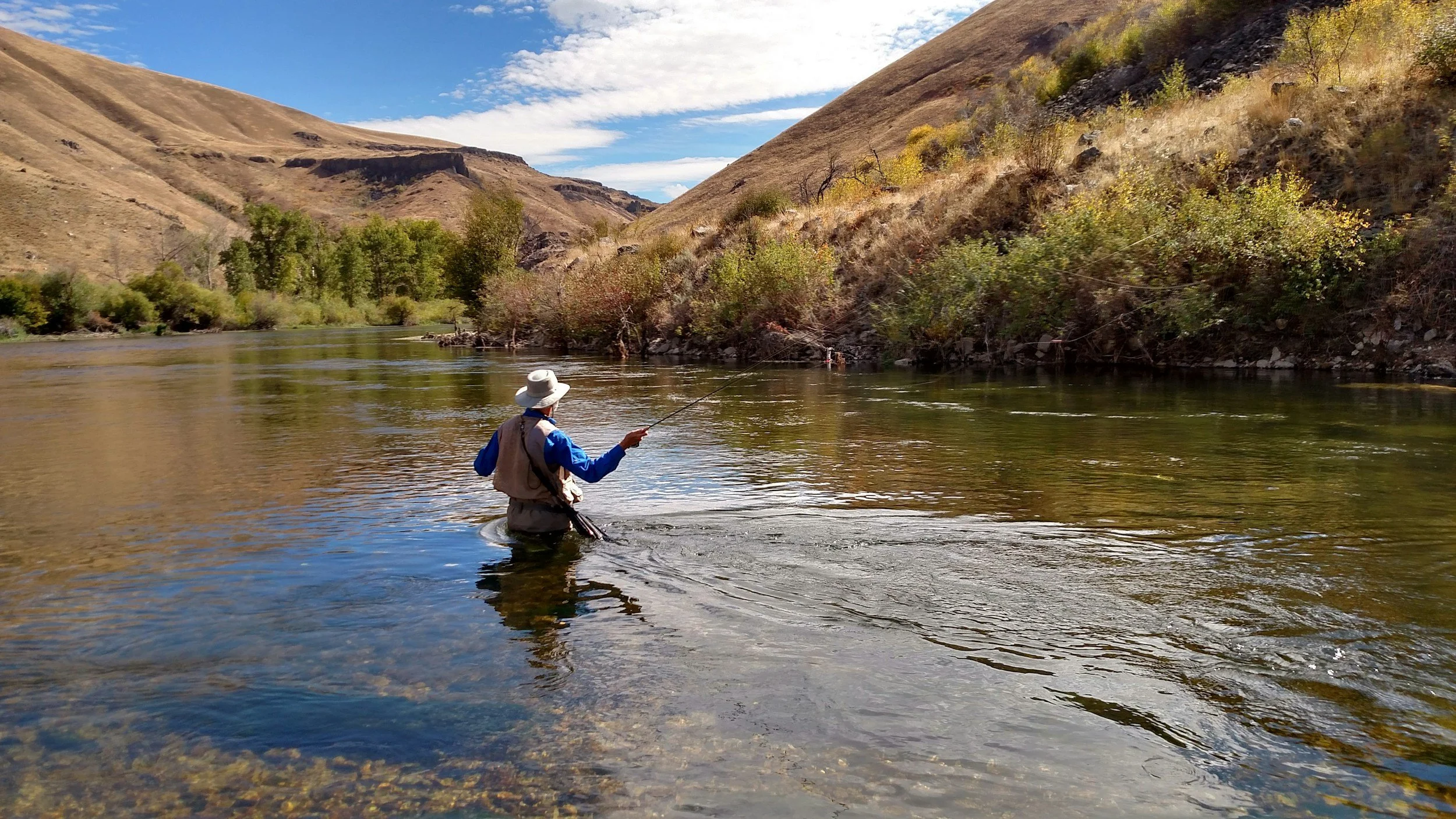 Person fishing in a river with hills and trees in the background on a clear day.