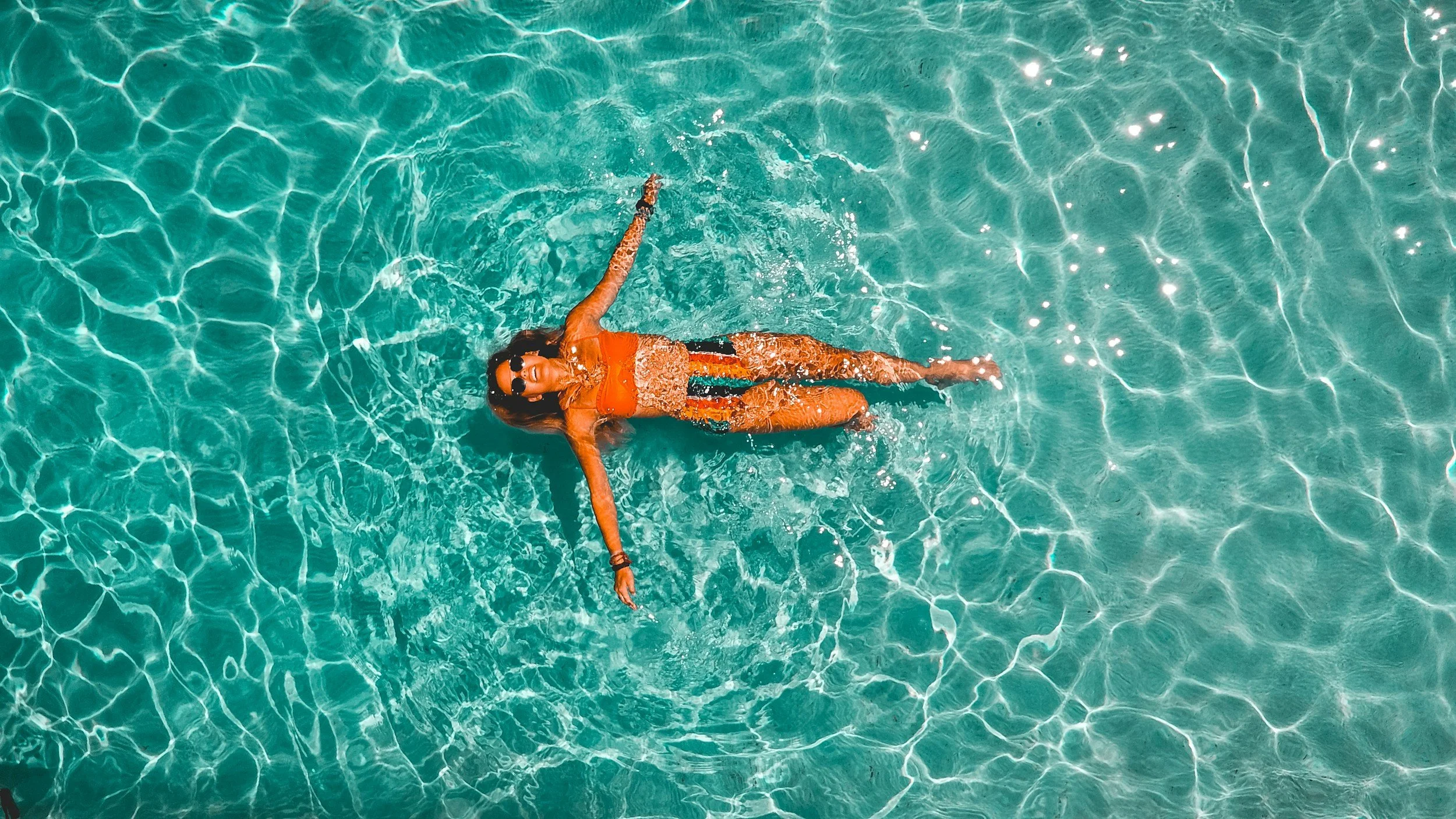 A woman in an orange bathing suit and black sunglasses floating in a clear blue swimming pool with sunlight reflecting off the water.