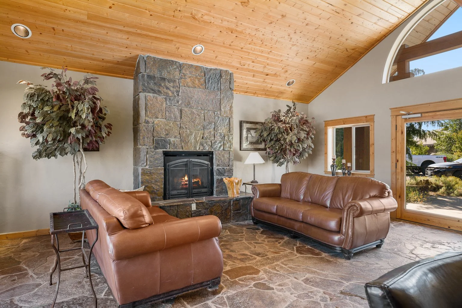 Cozy living room with leather sofas, stone fireplace, large window, and wood-paneled ceiling.