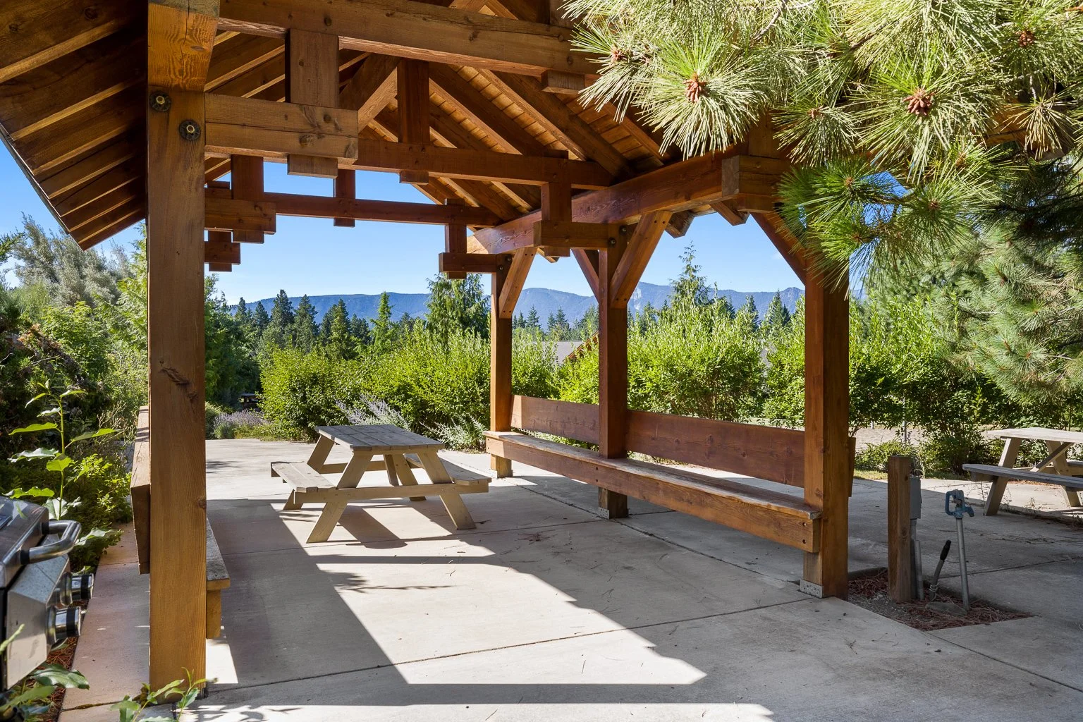 A wooden gazebo with a bench and a picnic table underneath, surrounded by greenery, with mountains in the distance.