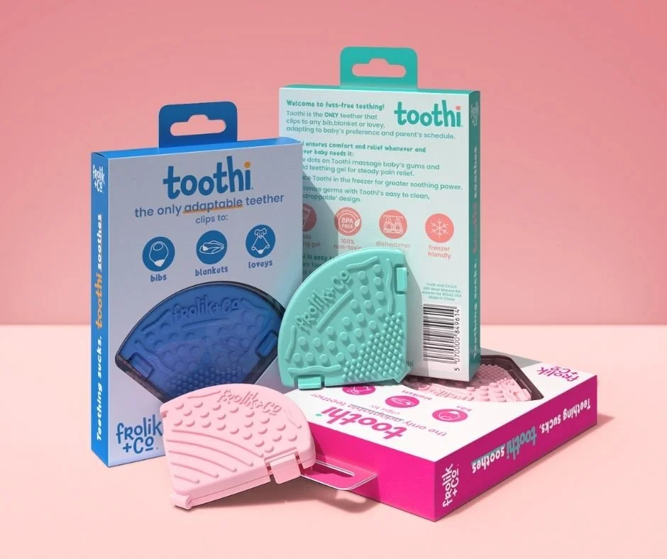 Collection of Toothi teething clips in pink, blue, and mint green, along with packaging and accessories, set against a pink background.
