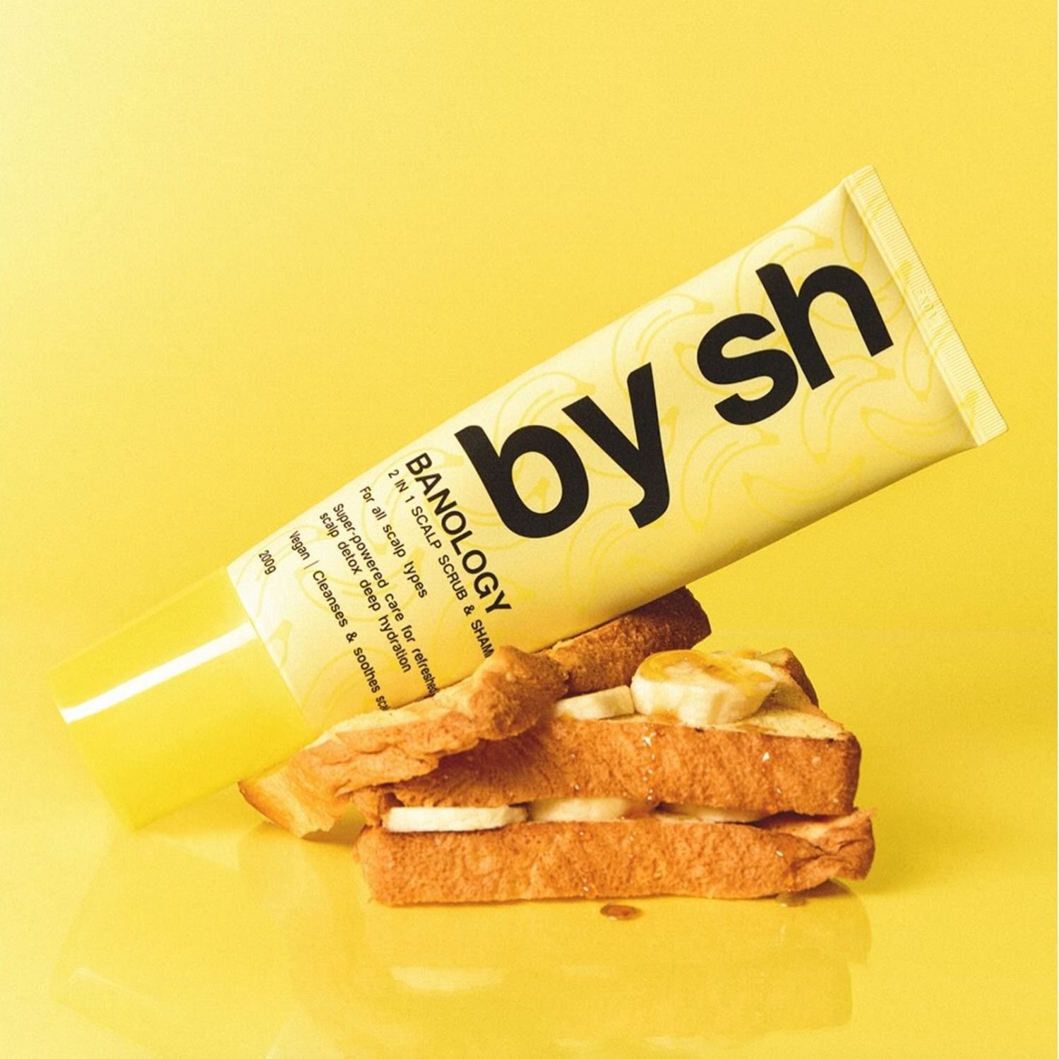 A tube of Biodology by SH herbal scalp scrub placed on top of a slice of toasted bread with banana slices on top, all set against a yellow background.