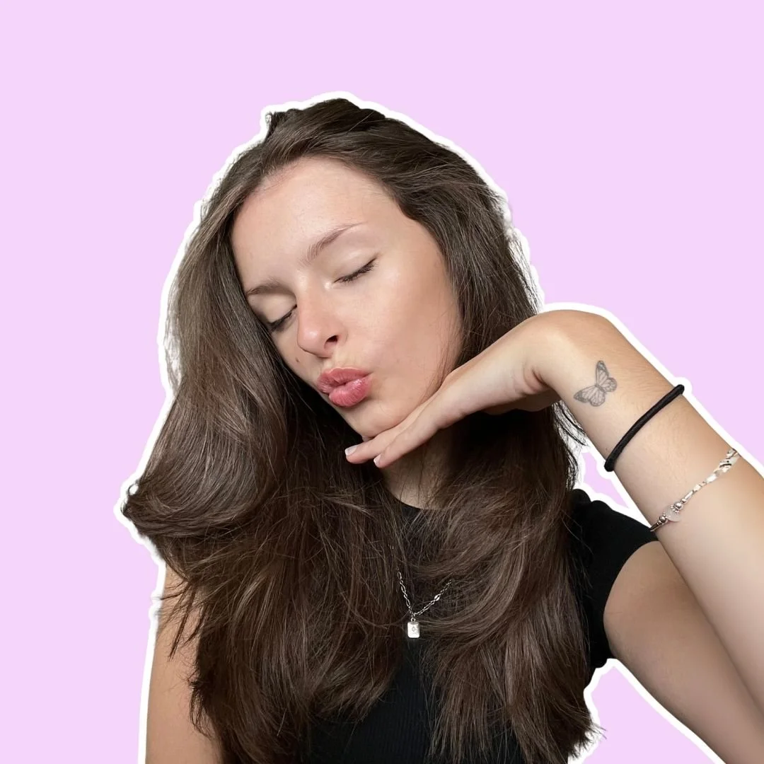 A young woman with long brown hair and closed eyes, making a kissing face and resting her chin on her hand, against a purple background.