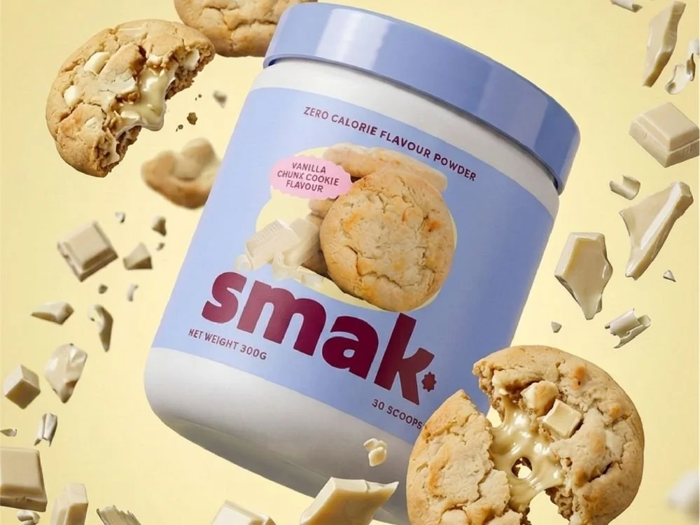 A container of small vanilla chunk cookie flavor powdered supplement surrounded by broken cookie pieces and white chocolate chunks on a light yellow background.