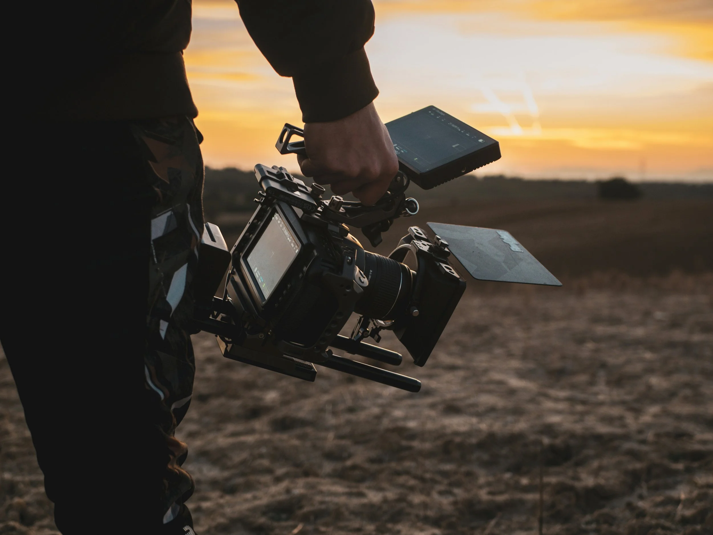 A person operating a drone or camera equipment near a body of water during sunset, with the focus on their hand holding the device and the camera setup.