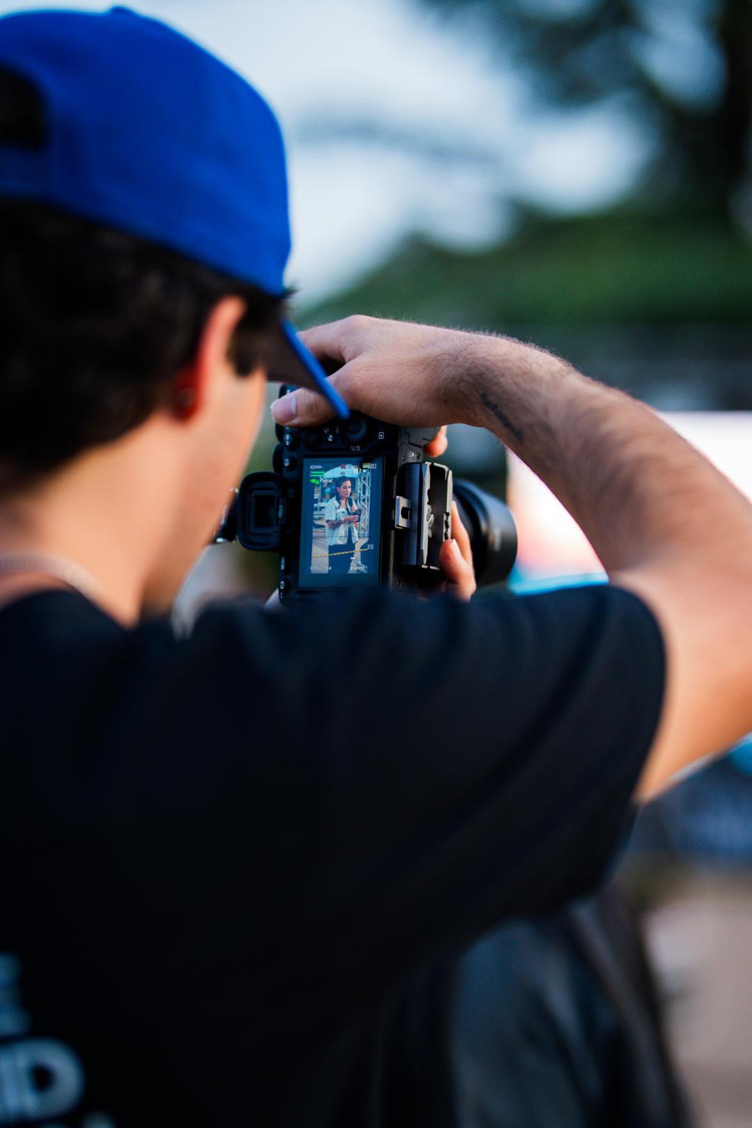 Person wearing a blue cap taking a photo with a professional camera, reflection of the person holding a phone visible in the camera's screen, outdoors during daytime.