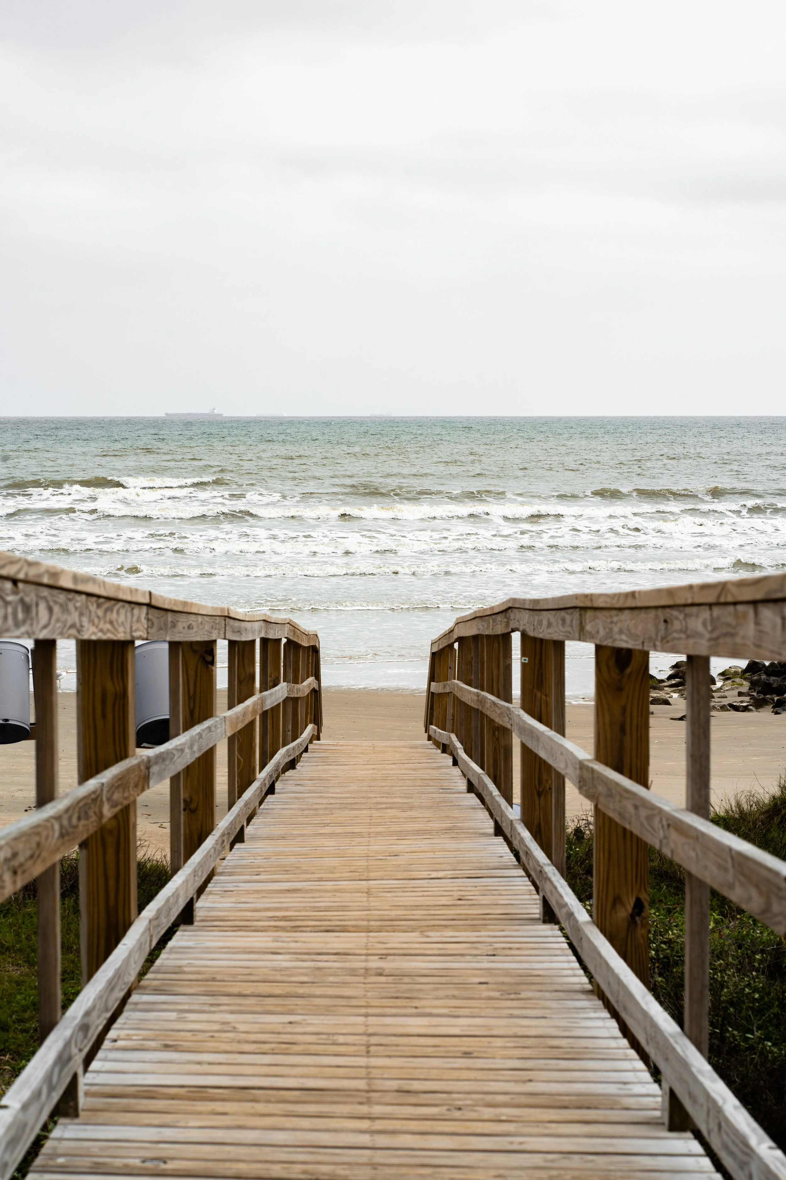 Wooden walkway leading to the beach with ocean waves and cloudy sky in the background.