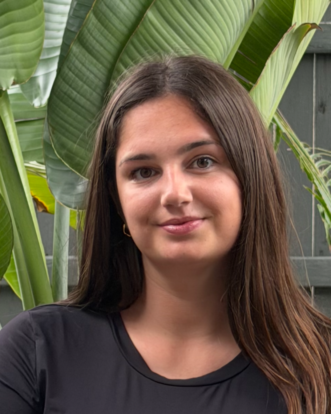A young woman with long brown hair, wearing a black shirt, standing in front of lush green tropical plants.