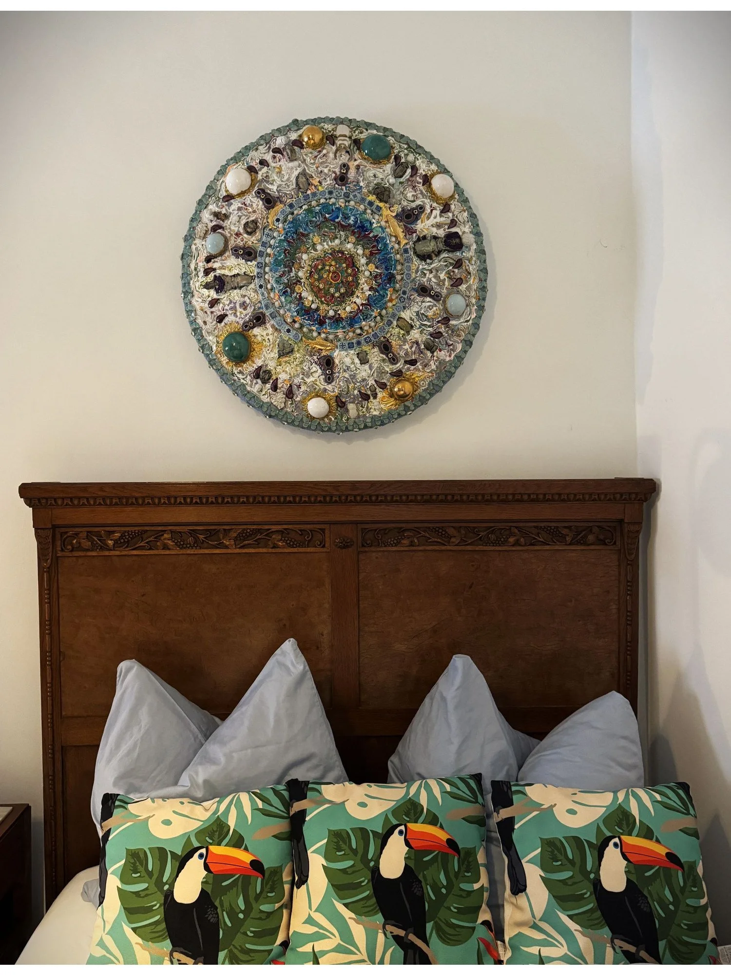 Gemütliches Schlafzimmer mit vier Kopfkissen und drei Dekokissen mit Tukan-Design auf einem holzgeschnitztem Holzbett. Über dem Bett hängt ein kunstvolles, rundes Wandbild aus bunten Perlen und andere Ornamente