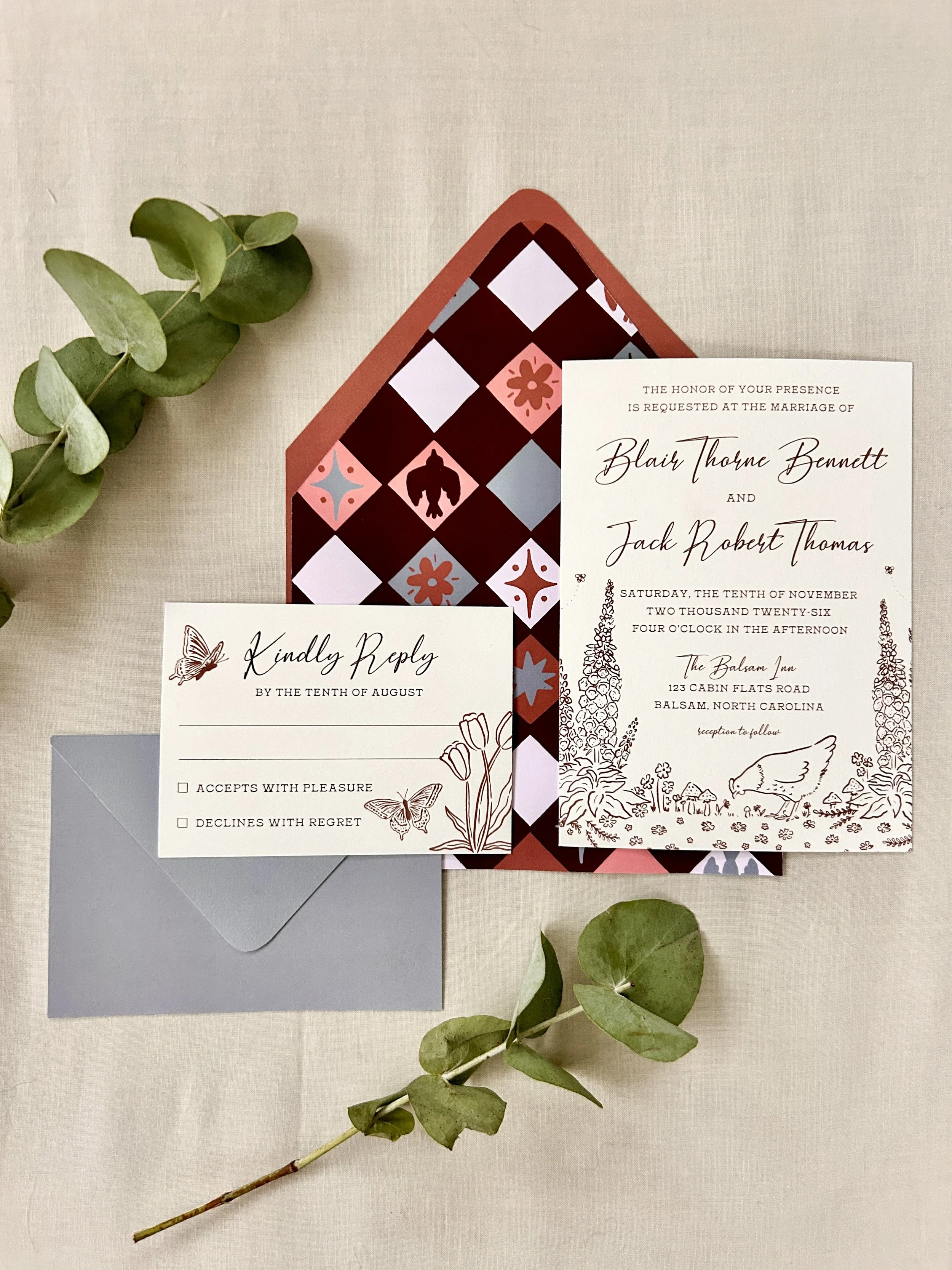 Wedding invitation, RSVP card, and envelope placed on a beige cloth surface with green eucalyptus leaves.