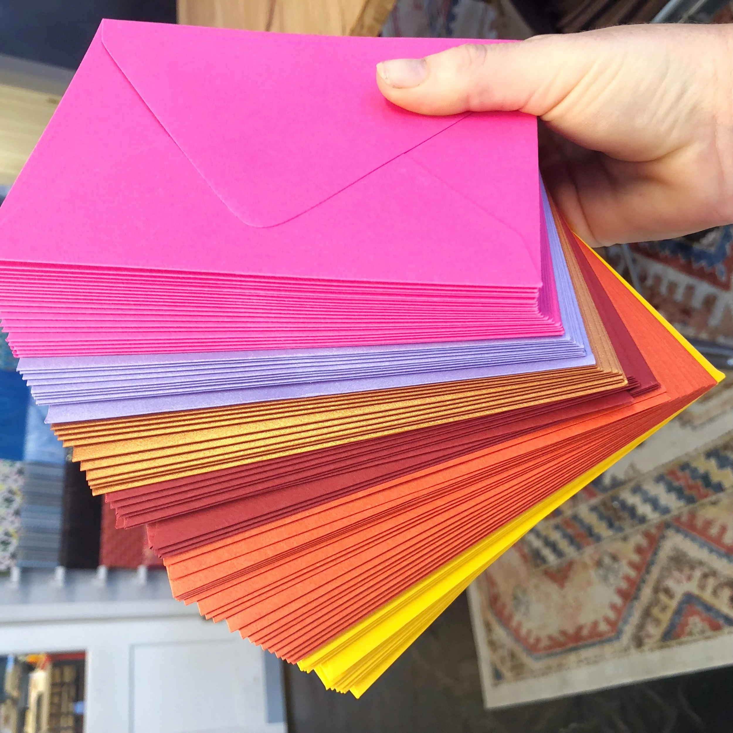 A hand holding colorful envelopes in shades of pink, purple, orange, and yellow, with a patterned rug and shelves in the background.