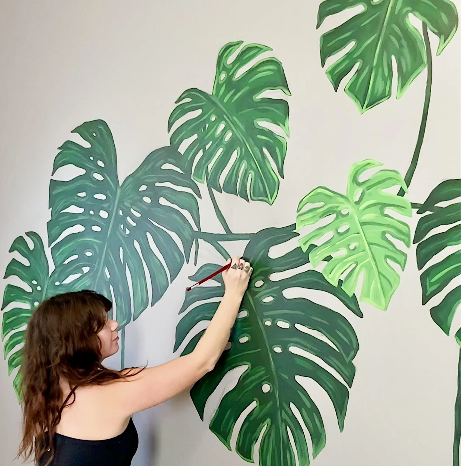 A woman painting large green Monstera plant leaves on a wall.