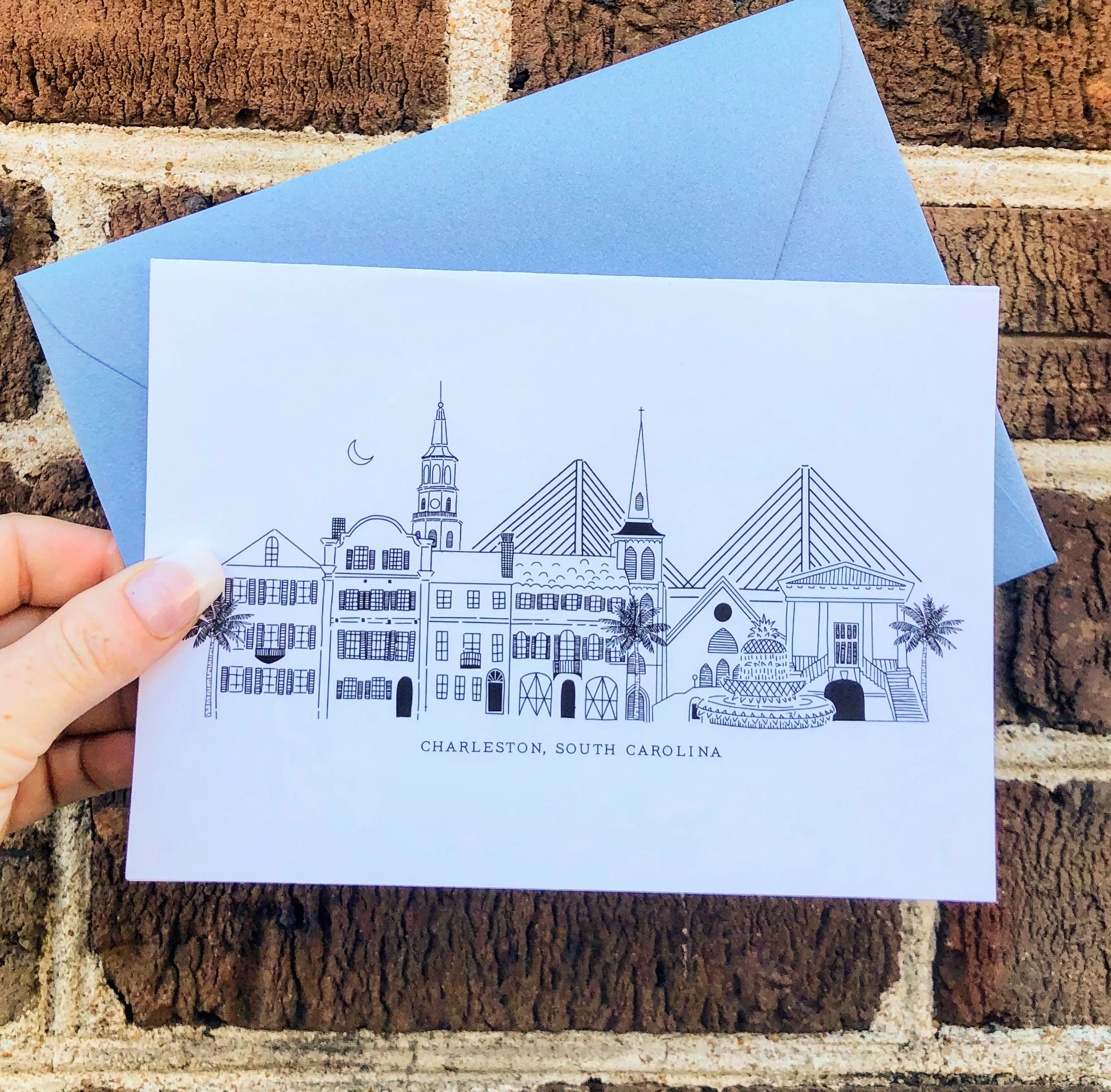 A hand holding a postcard with a line drawing of Charleston, South Carolina's skyline, featuring historical buildings, palm trees, and bridges, with a crescent moon in the sky and the text 'CHARLESTON, SOUTH CAROLINA'.