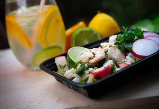 A black tray of seafood salad with sliced radishes and lime wedges. A glass of lemon-infused water with lemon slices and a straw is in the background, along with whole lemons.