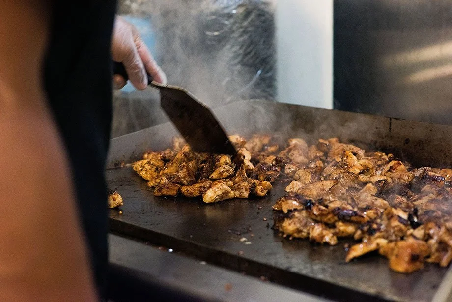 Close-up of seasoned chicken sizzling on a flat-top grill as a cook turns the pieces with a metal spatula, steam rising from the hot surface.