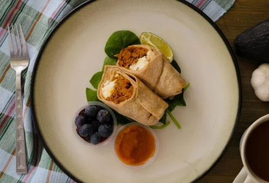 Two breakfast burritos on a plate with lime wedge, greens, blueberries, and sauce, with a fork on the left side.