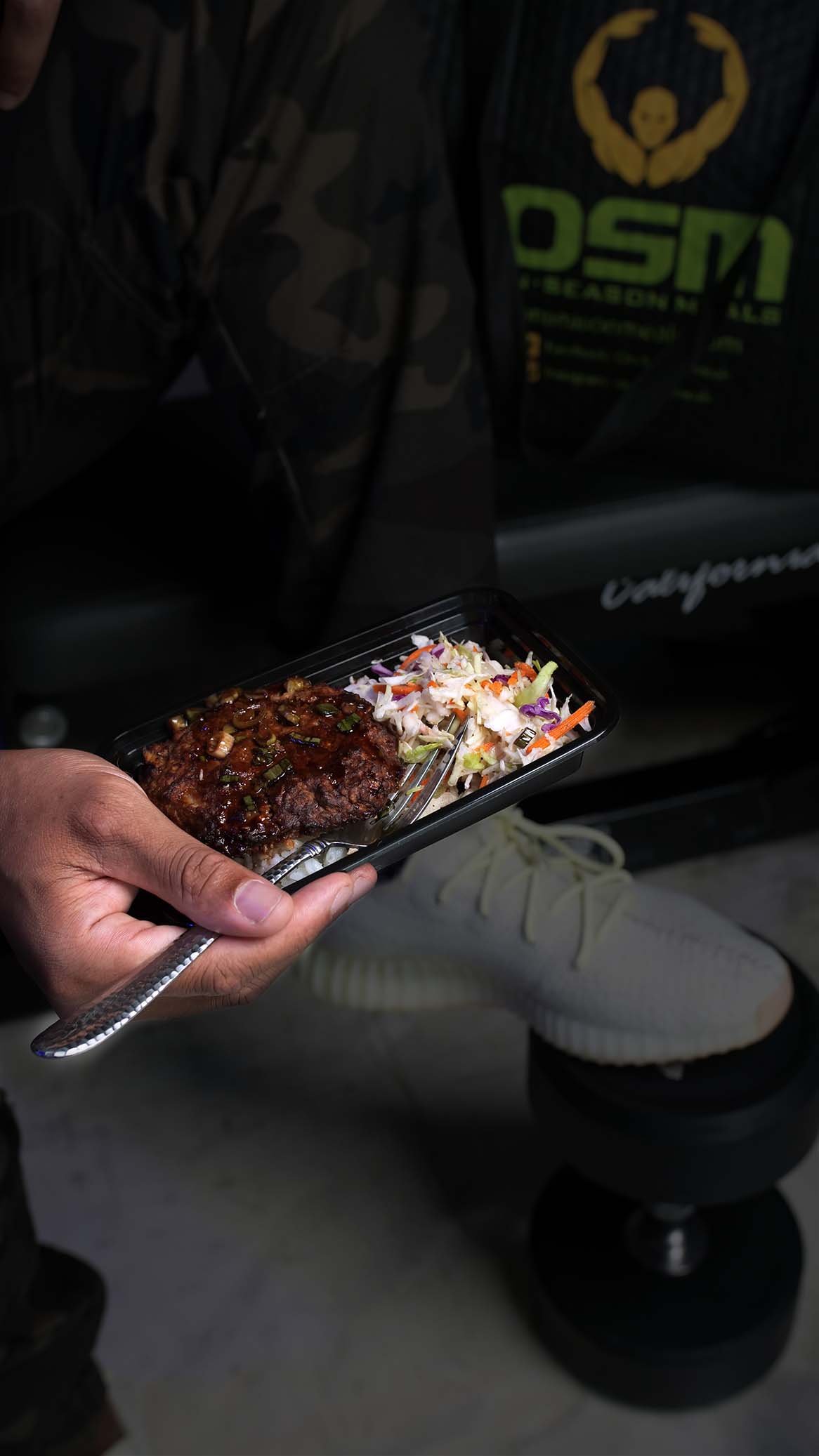 Hand holding a black takeout container with grilled beef over rice and fresh coleslaw, representing a convenient, ready-to-eat street food meal in a casual urban setting.