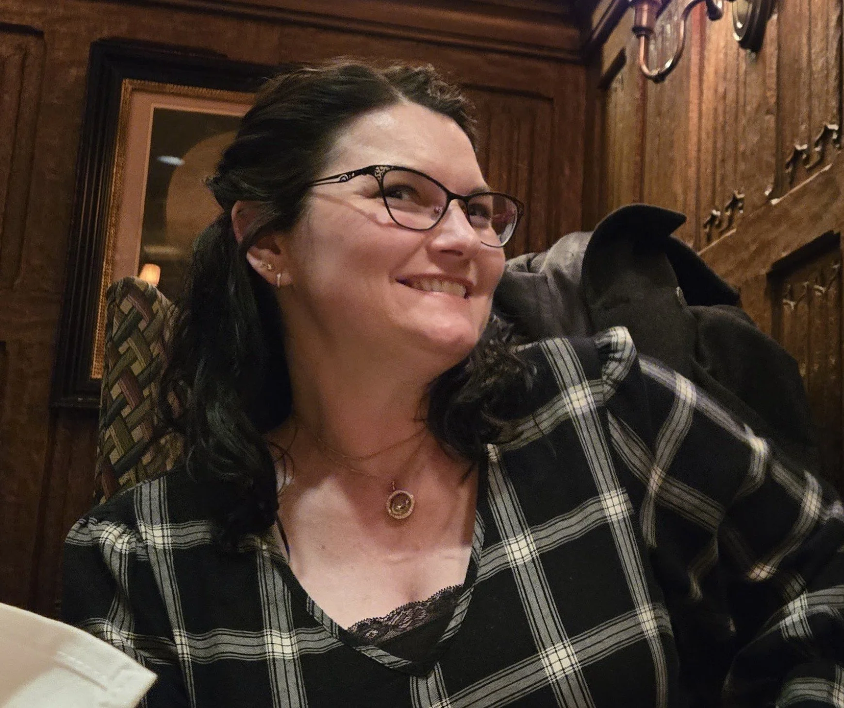 A woman with glasses smiling, wearing a plaid shirt and jewelry, sitting in a wood-paneled restaurant or room with decorative wall details.