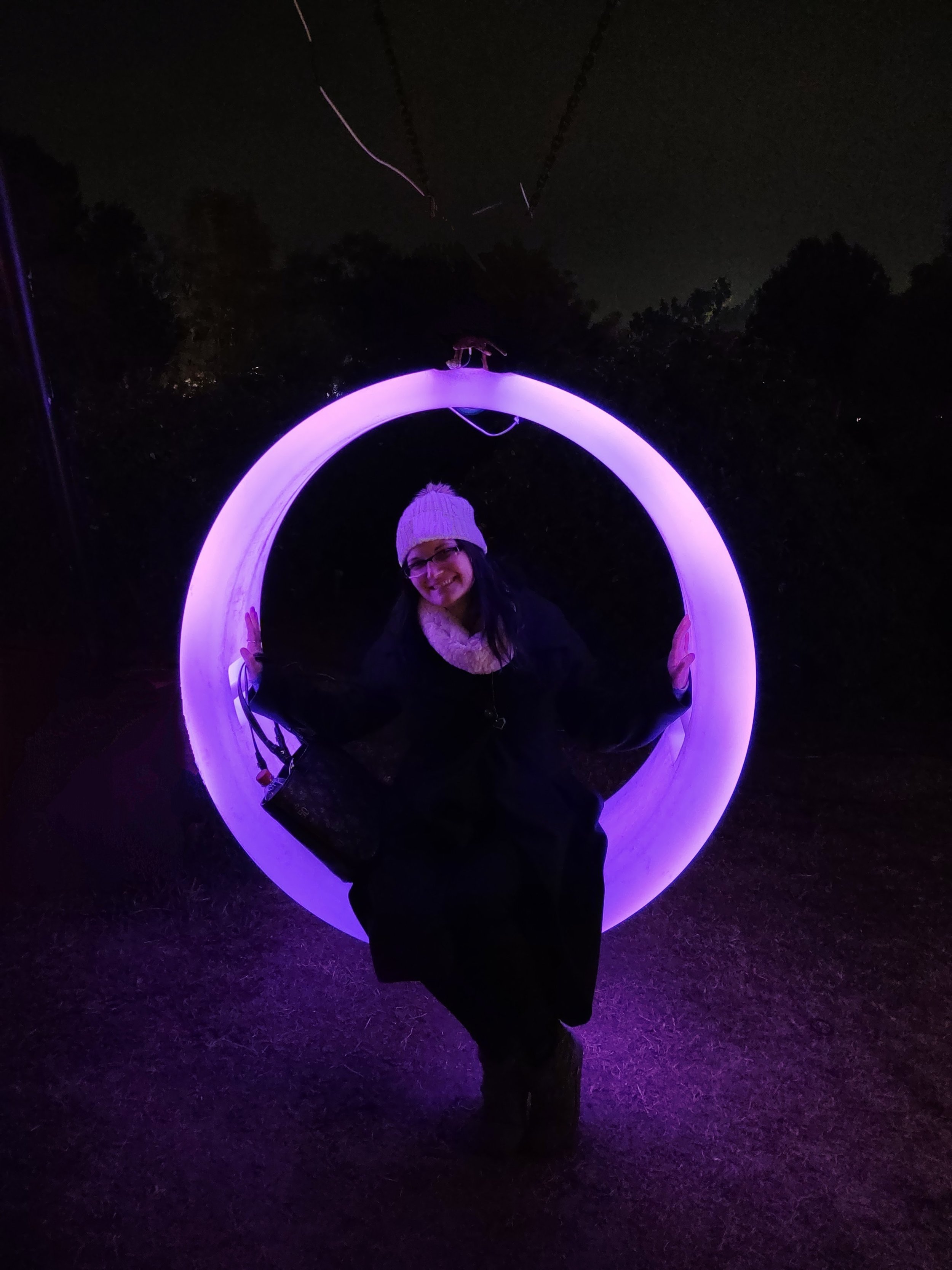 Person smiling and sitting inside illuminated purple ring at night outdoors, wearing a hat and dark clothing.