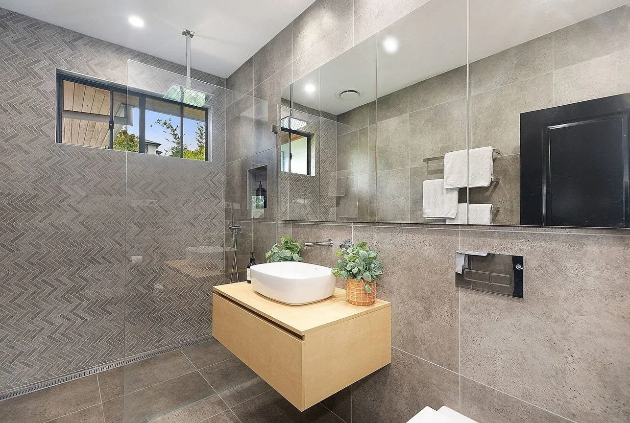 Modern bathroom with gray tiled walls and a large mirror, featuring a white vessel sink on a wooden floating vanity, with green plants and towels visible in the reflection.