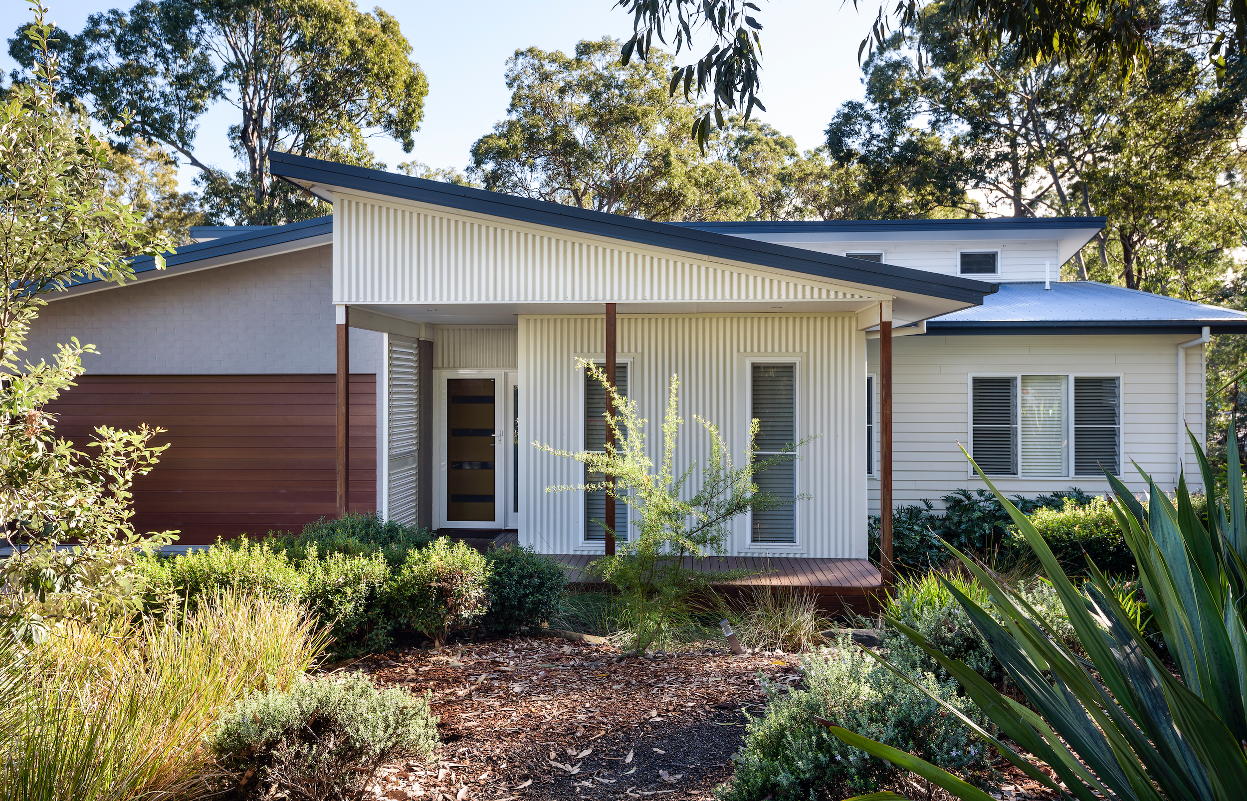 Modern house with a mix of white siding and wood accents, surrounded by bushes and trees, with a garden and pathway in front.