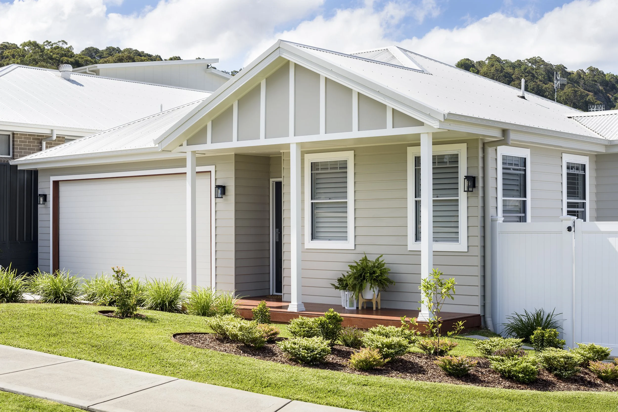 A modern suburban house with beige siding, white trim, and a white metal roof. The house has a small front porch with potted plants and black outdoor wall lanterns. Well-maintained garden with trimmed bushes and grass lines the sidewalk.