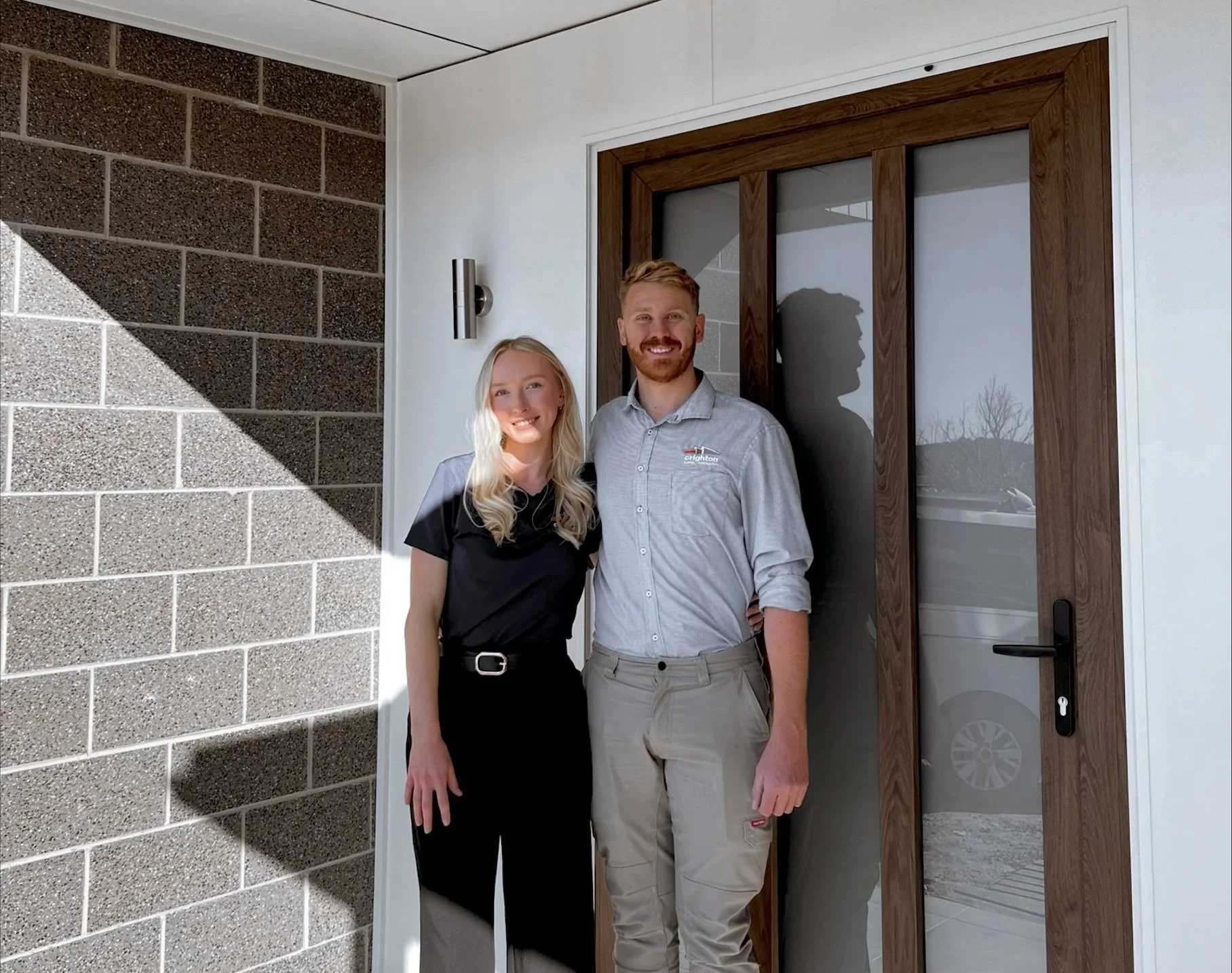 A smiling man and woman standing together outside a house, near a wooden-framed glass front door, with the man wearing a light grey shirt and beige pants, and the woman in a black top and black pants, with a brick wall on the left and a white wall on the right.