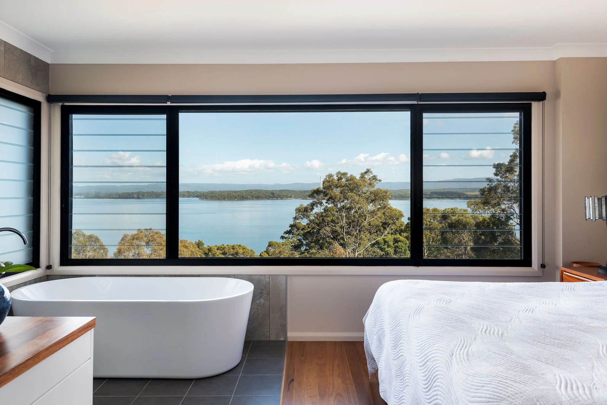 Bedroom with large window viewing a lake, with a white bathtub near the window and a bed with white linens.