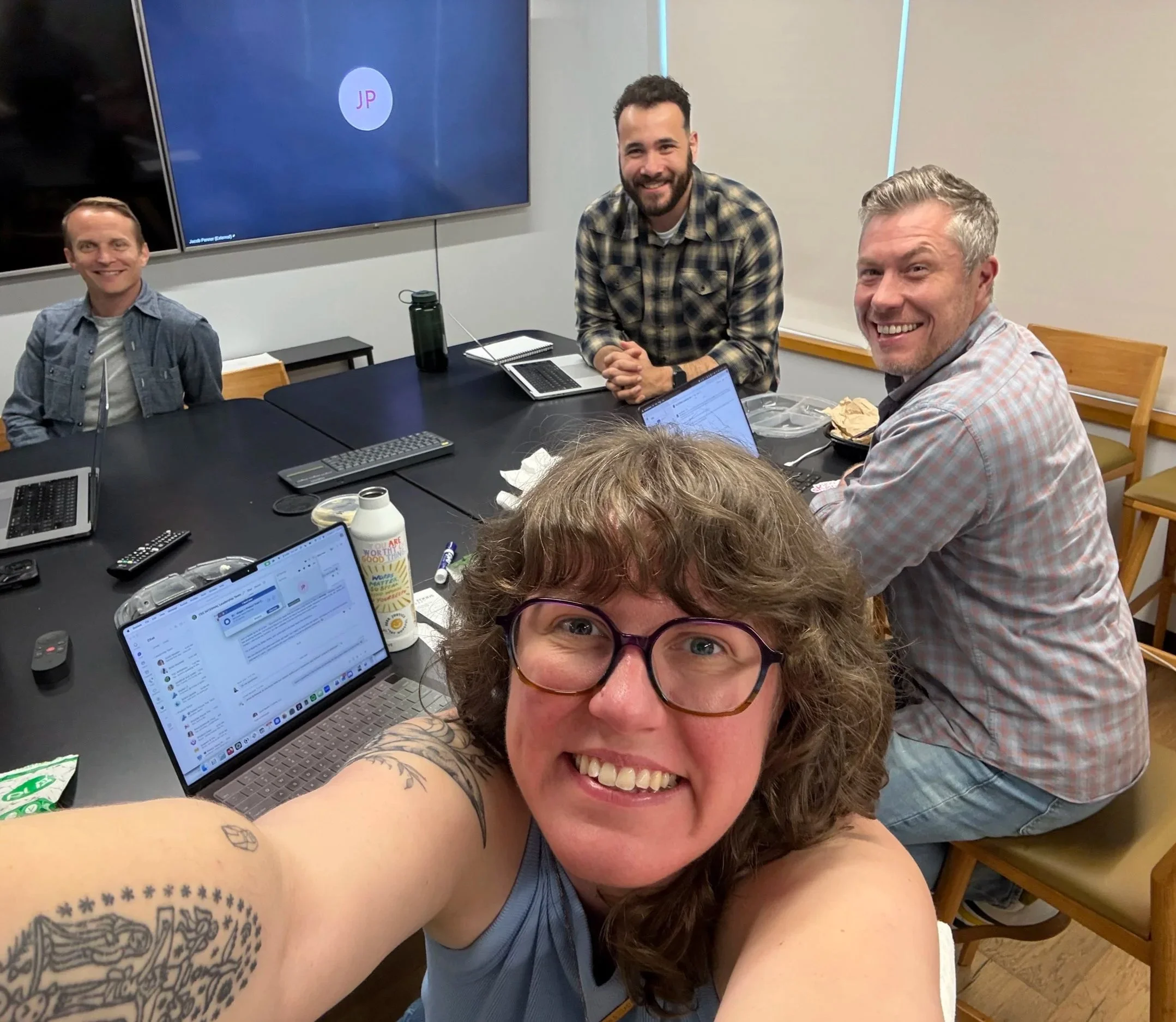 A group of five people in a meeting room, smiling for a selfie, with laptops, notebooks, and drinks on the table.