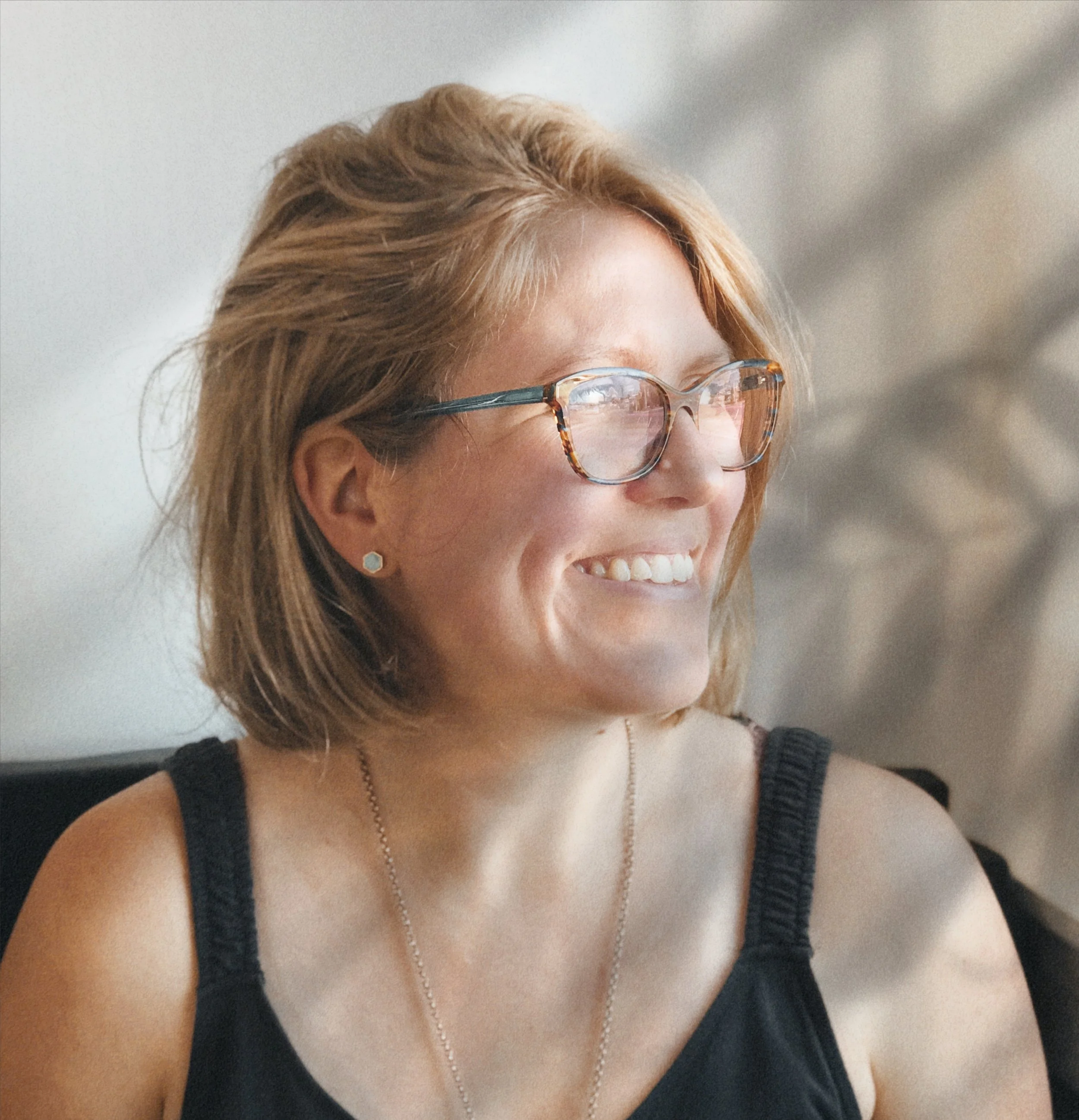 A smiling woman with short, light brown hair, wearing glasses with tortoise shell frames, a black tank top, small earrings, and a delicate necklace, sitting indoors.
