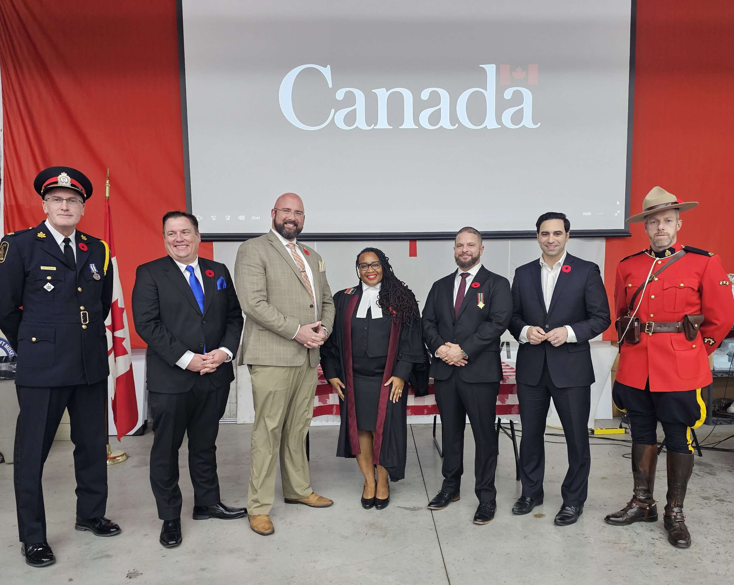 Seven individuals in formal and historic military attire, standing in front of a large screen displaying the word 'Canada' with a small Canadian flag icon, during a formal event or ceremony. They are posing for a group photo.