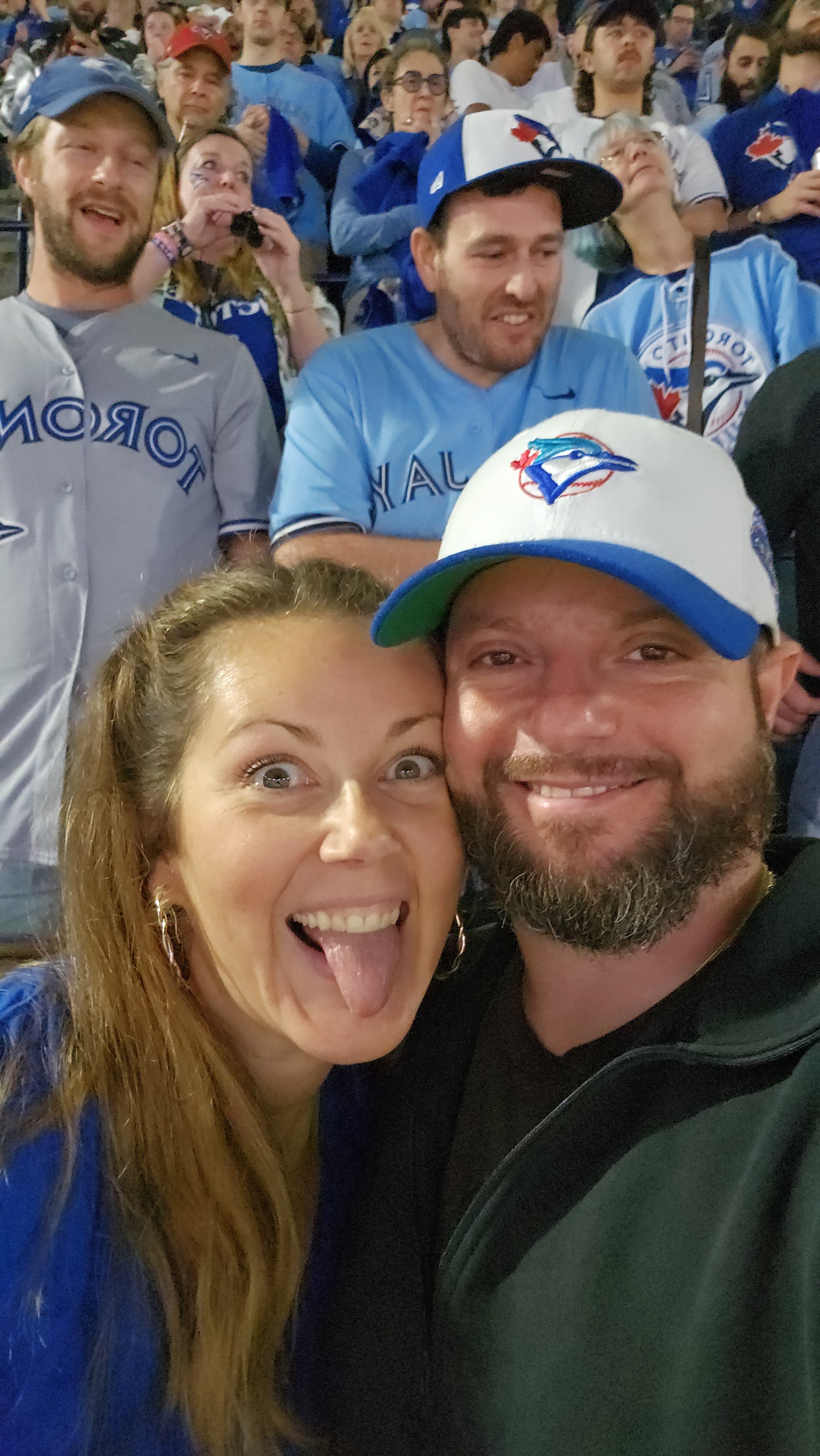 A group of Toronto Blue Jays fans at a game, with two people in the foreground taking a selfie. The woman is sticking out her tongue and smiling, while the man is smiling and wearing a Blue Jays hat. The crowd behind them is also wearing Blue Jays merchandise and appears excited.