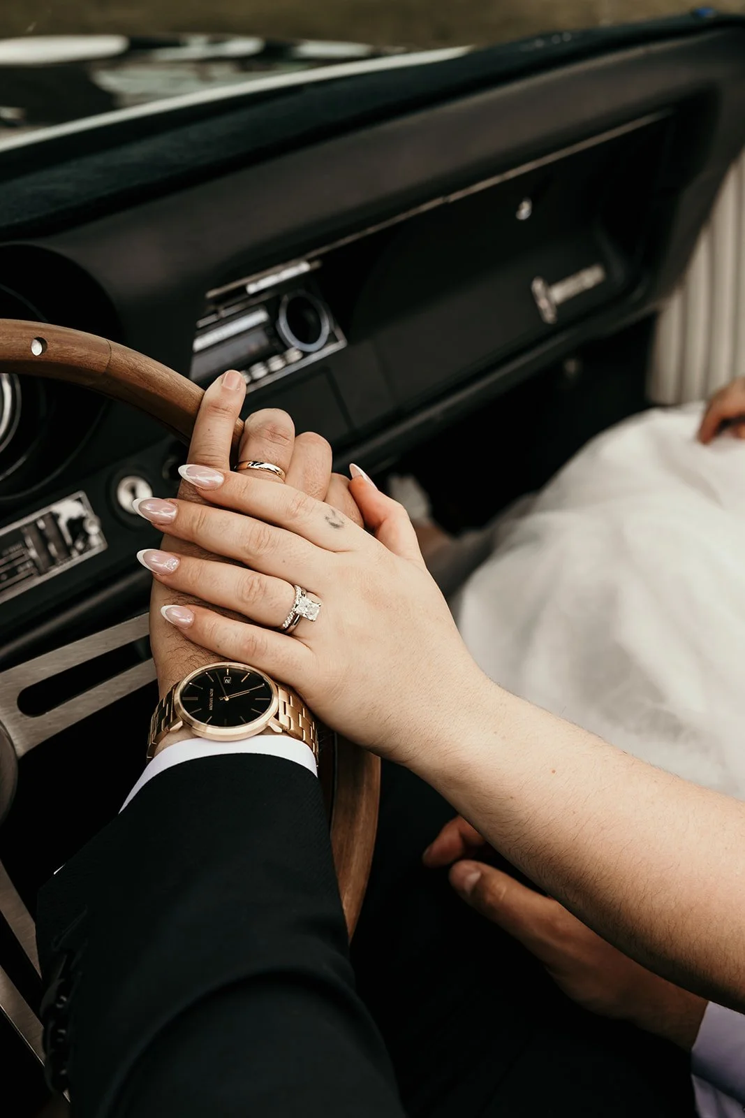 Close-up of a couple's hands holding each other on a vintage car's steering wheel. The woman has manicured nails and a diamond ring, while the man wears a gold watch and a dark suit.