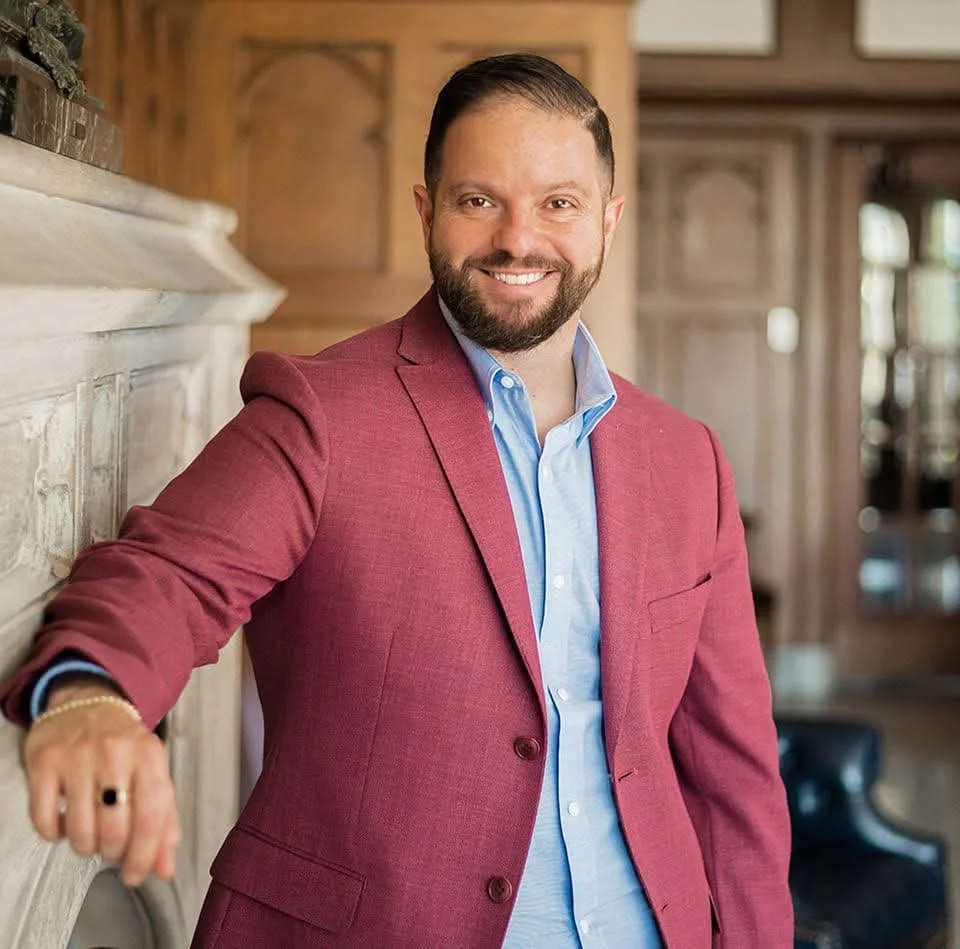 A man with a beard and short dark hair, wearing a red blazer and a light blue shirt, smiling while standing indoors against a background of wood paneling and furniture.