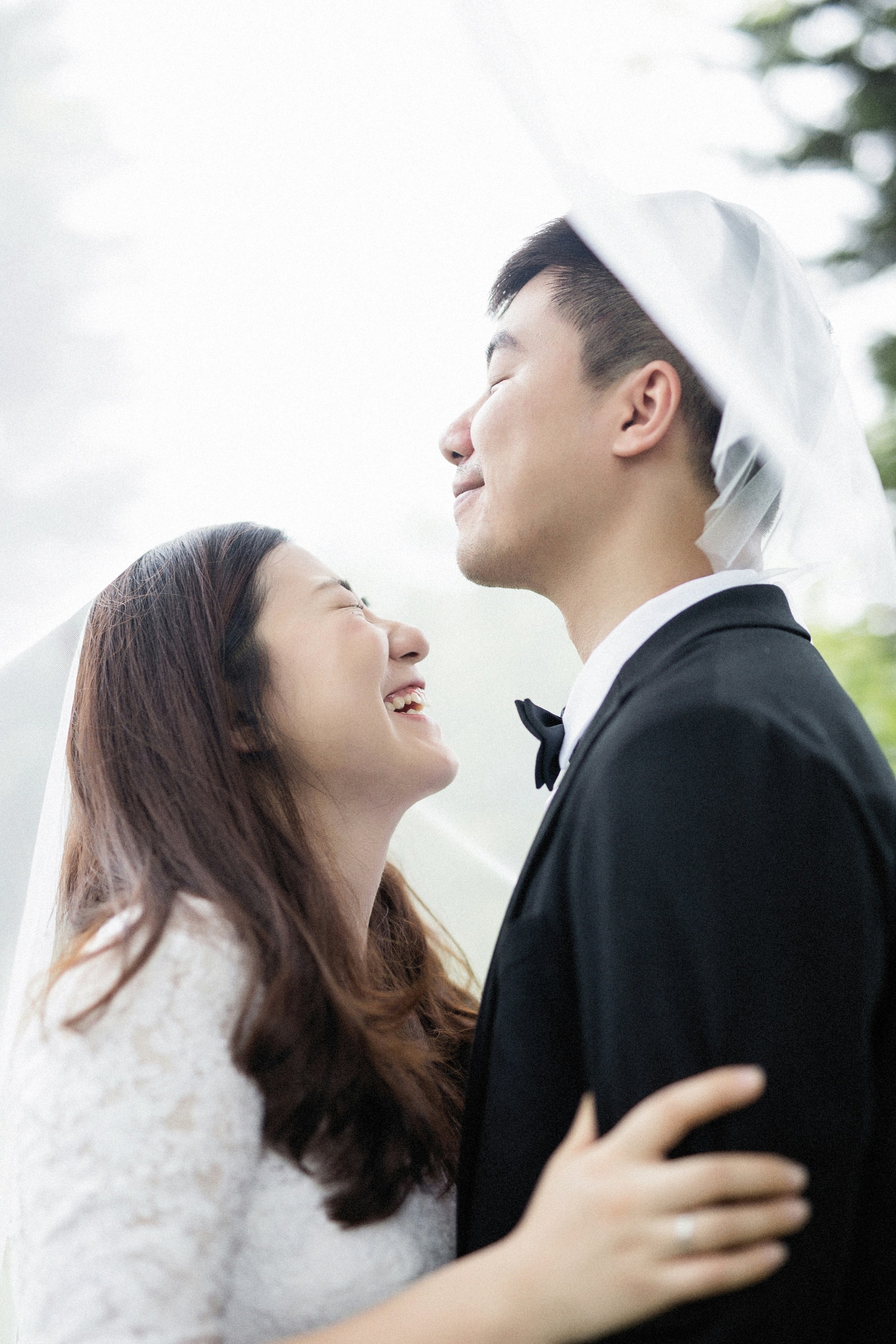 A newlywed couple sharing a moment outdoors, with the groom whispering to the bride, both smiling. The bride wears a lace wedding dress and a floral crown, while the groom is in a dark suit.