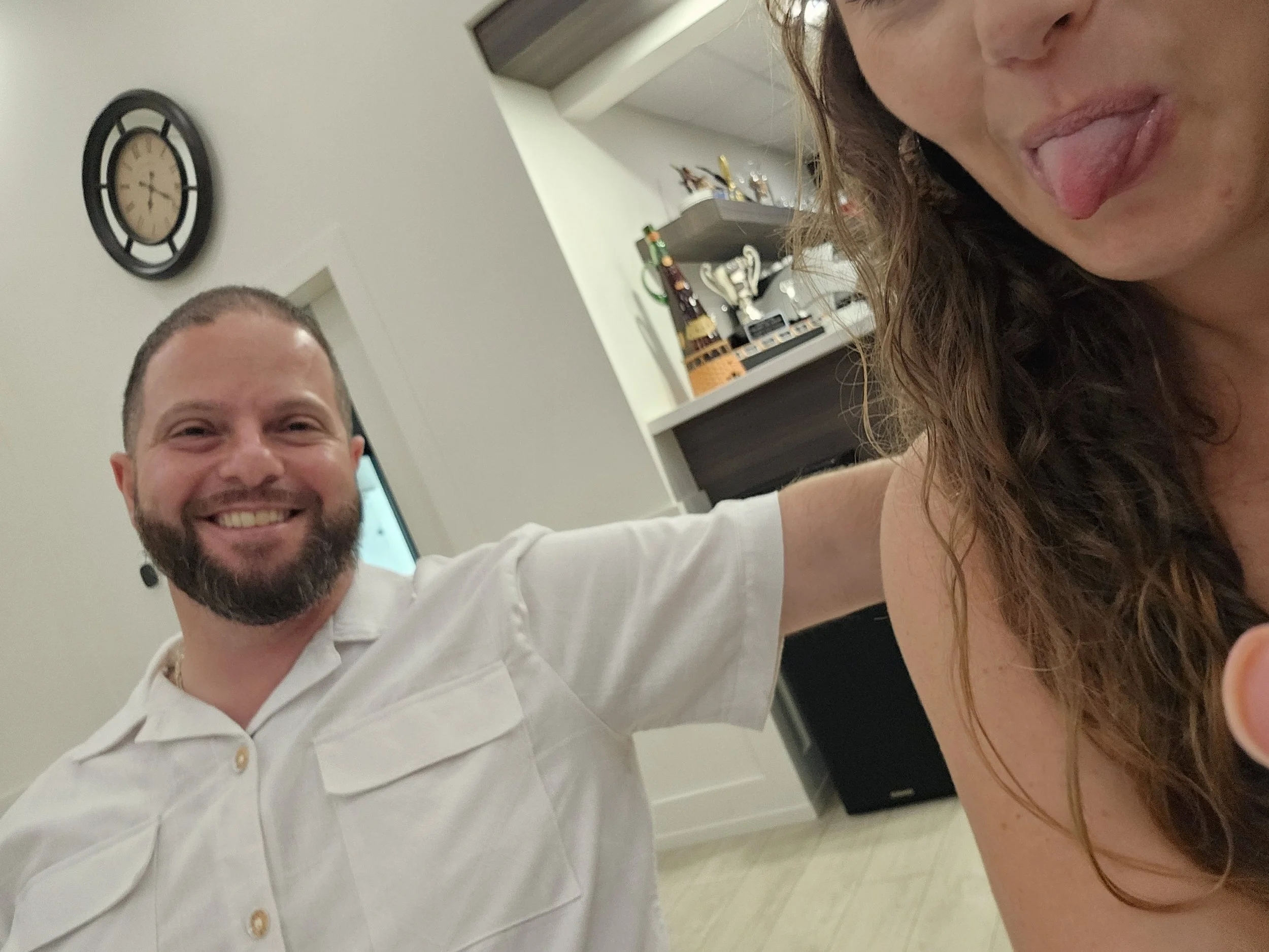 Smiling man with a beard taking a selfie with woman sticking out her tongue in a casual indoor setting.