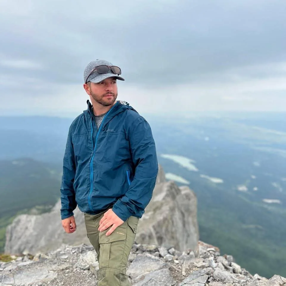 A man standing on a mountain summit with a vast landscape and cloudy sky in the background.