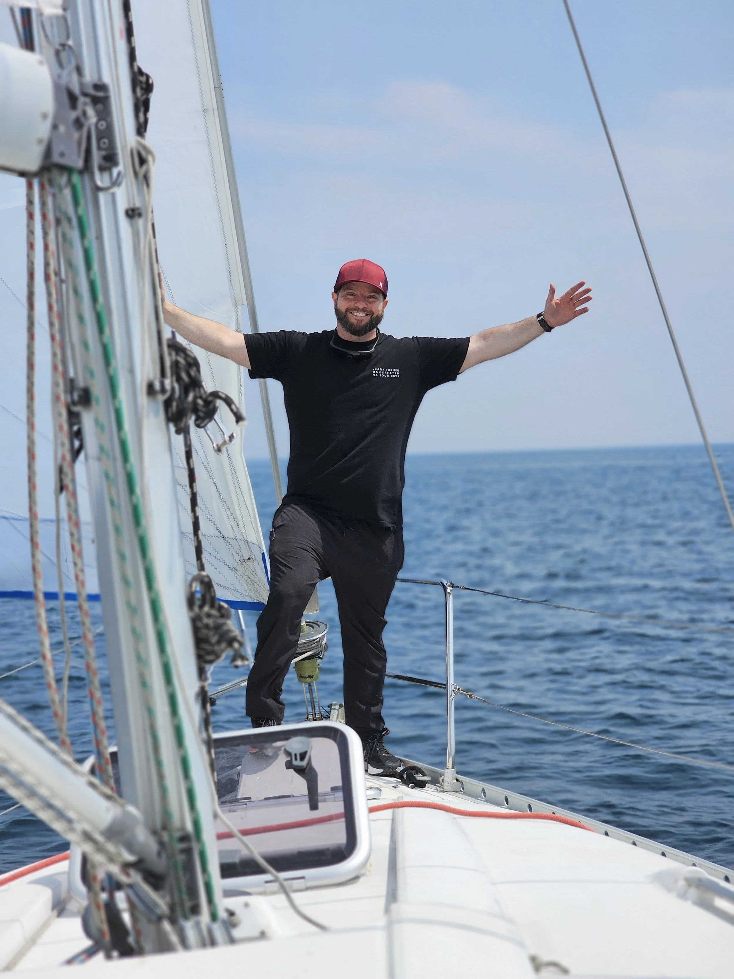 A man standing on a sailboat deck with arms outstretched, smiling, amid open water under a blue sky.