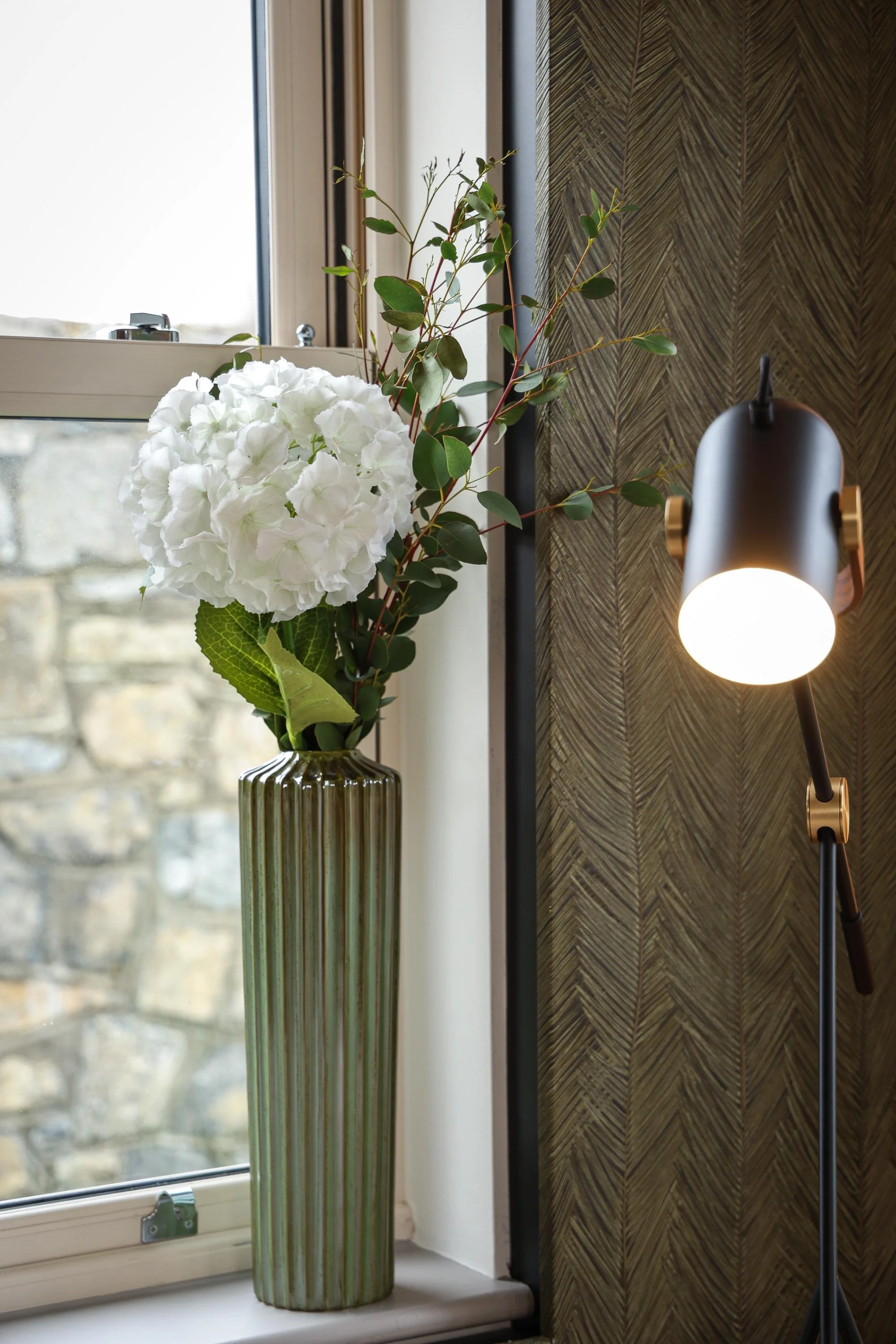 A tall, green, striped ceramic vase with white hydrangea flowers and green foliage on a windowsill, adjacent to a brown textured wall and a black lamp with a round white bulb.
